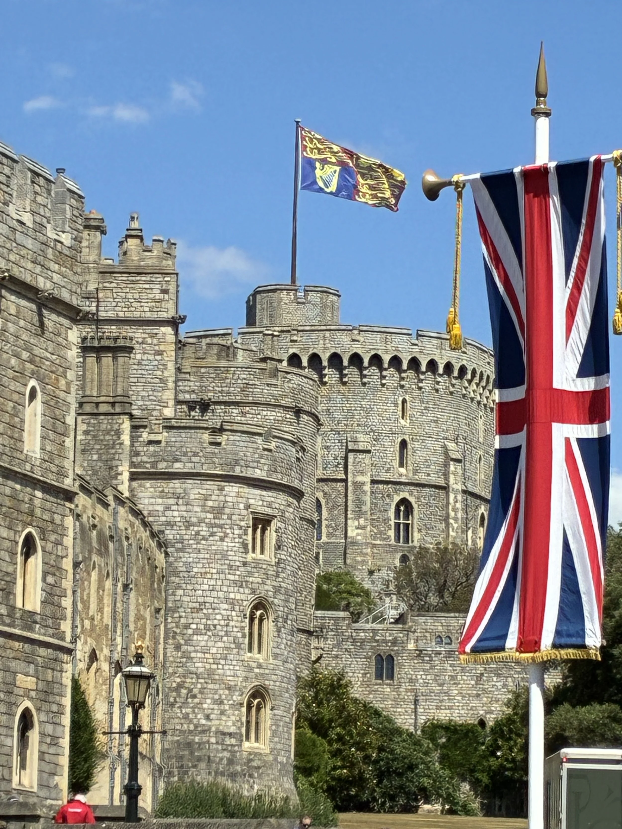 Windsor Castle , flanked by flagpoles with the Royal Standard and Union Jack flags. The sky is blue with a few clouds, and there are trees and a lamppost in the scene. We pass by here on our Windsor self-guided tour.