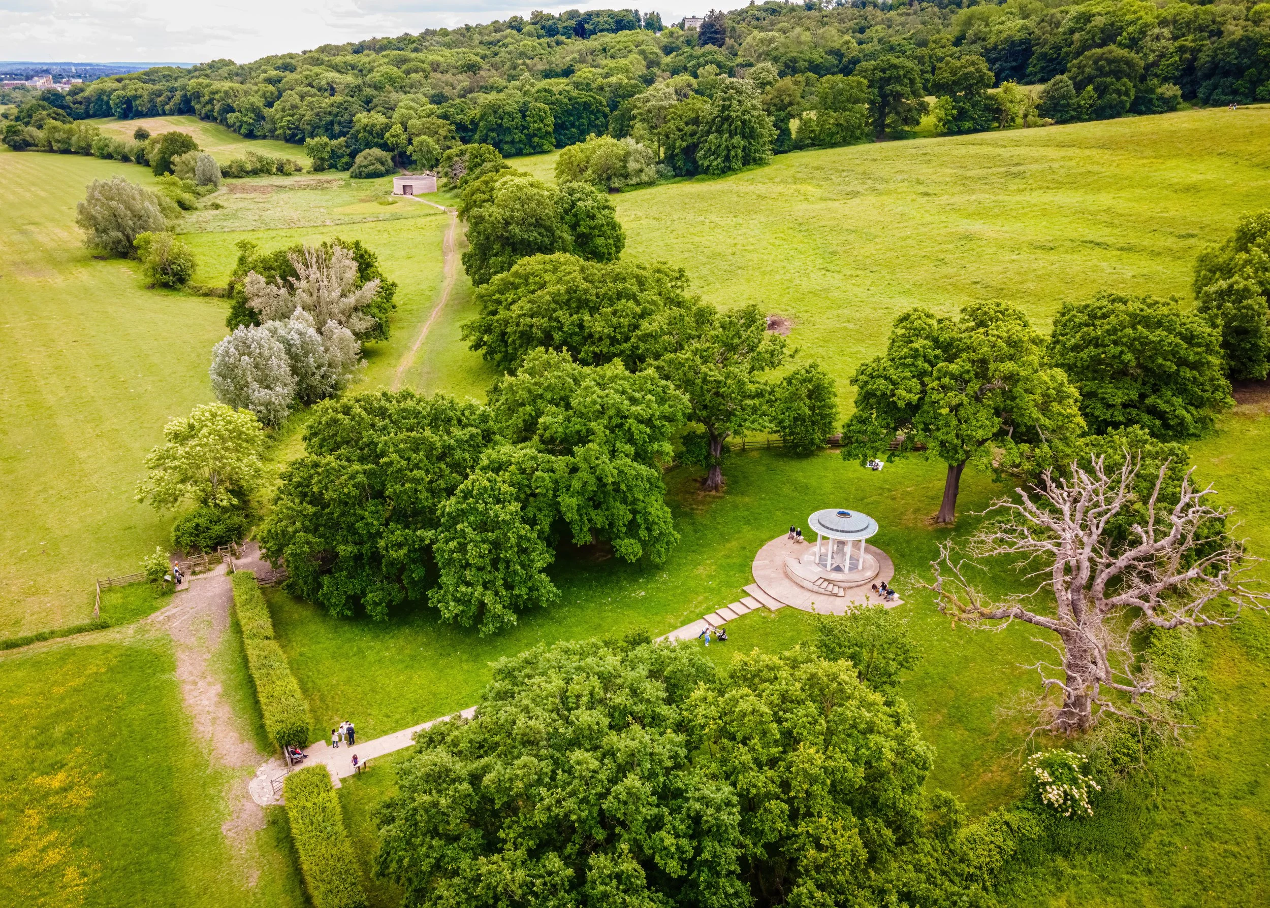 Aerial view of a park with lush green trees, a circular pavilion, paths, and people enjoying the outdoor space.