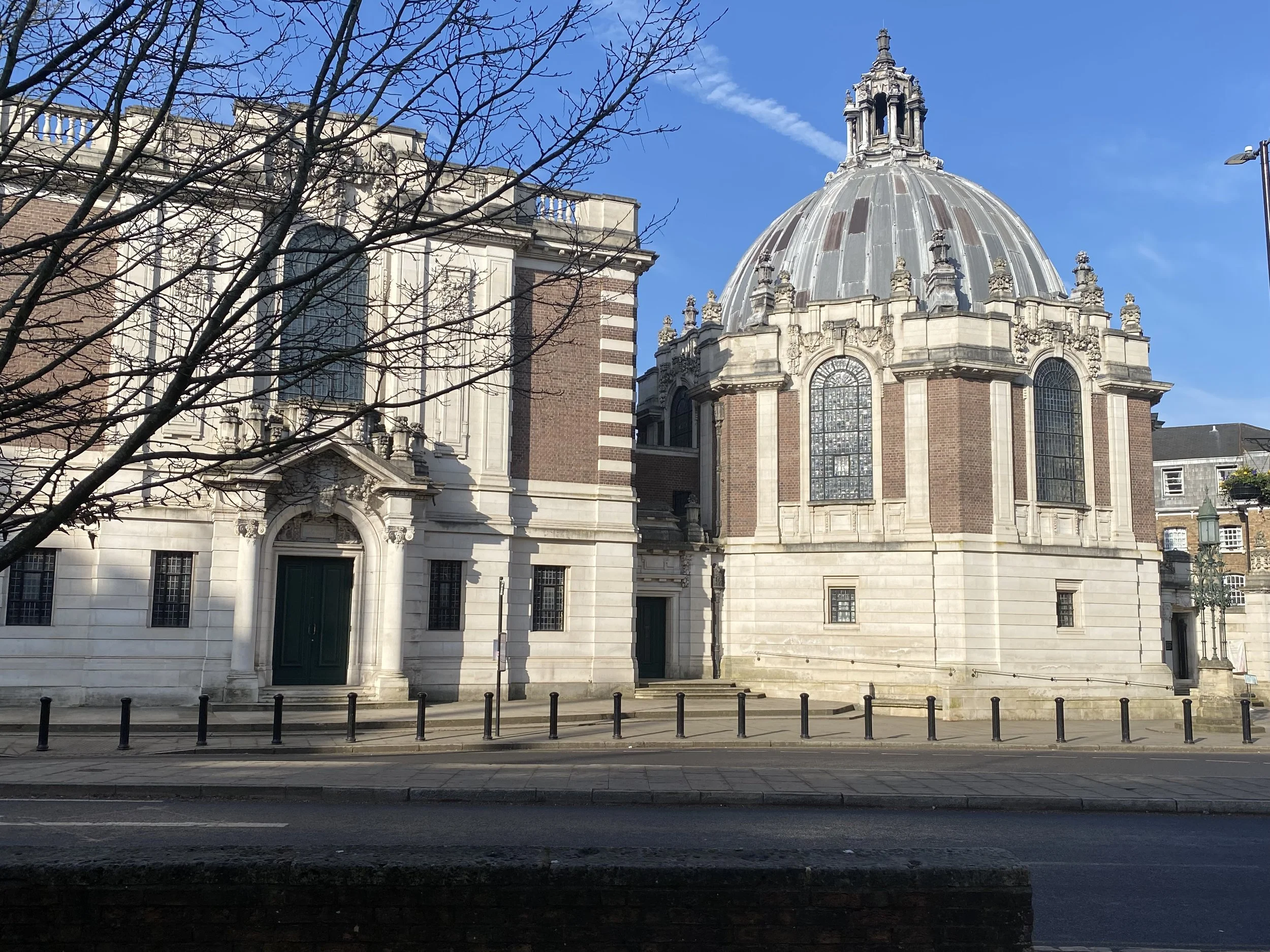 A historic building with a dome, brick and white stone architecture, and leafless trees in the foreground, under a clear blue sky.