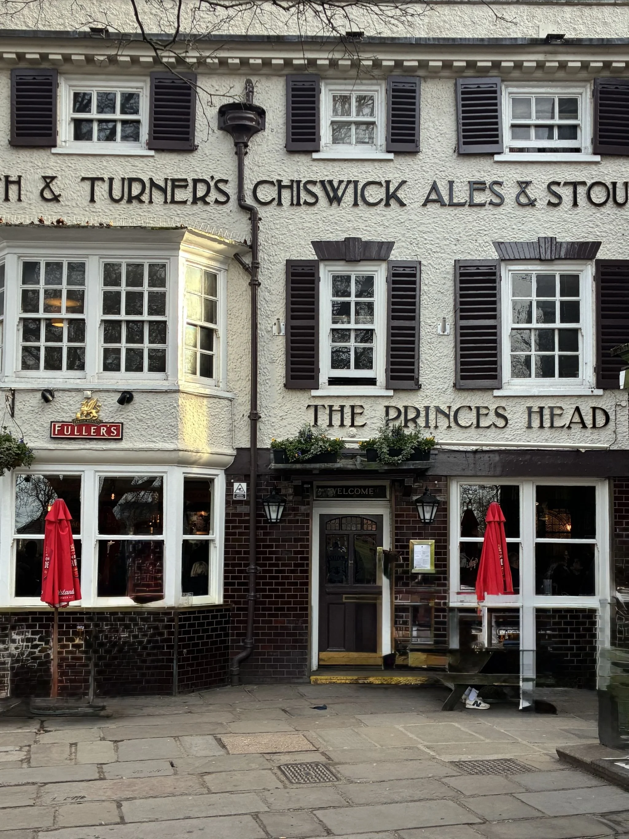 Exterior of a pub or restaurant building with black window shutters, white textured walls, and multiple windows. There are red umbrellas outside, and a sign for Fuller’s beer. The building has a sign with partial text reading, 'H & TURNER'S CHISWICK ALLES & STOU...' There are also black lantern-style lights and some plants in window boxes.