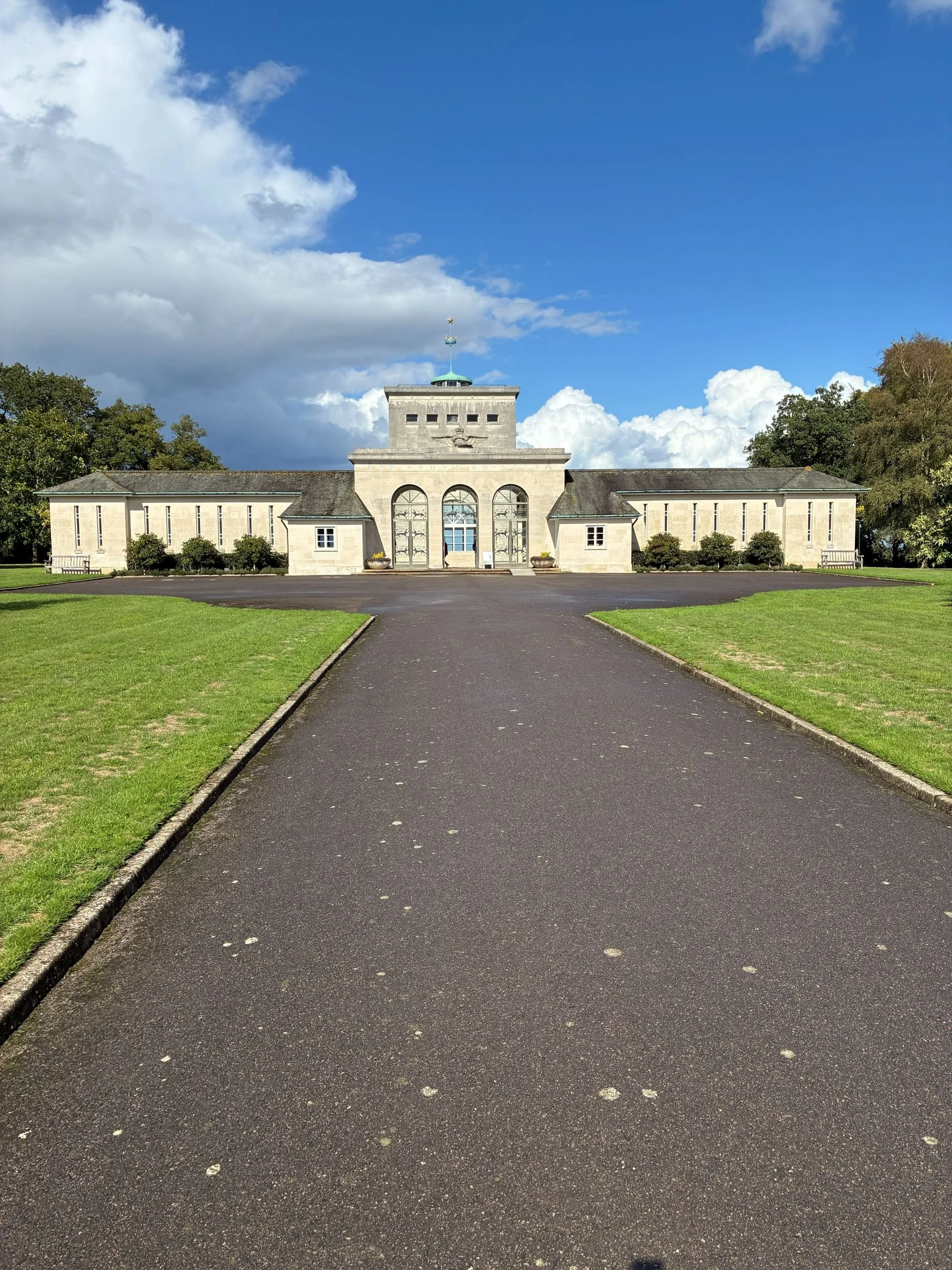 A historic building with arched windows and a central entrance, surrounded by green grass and trees under a partly cloudy sky.