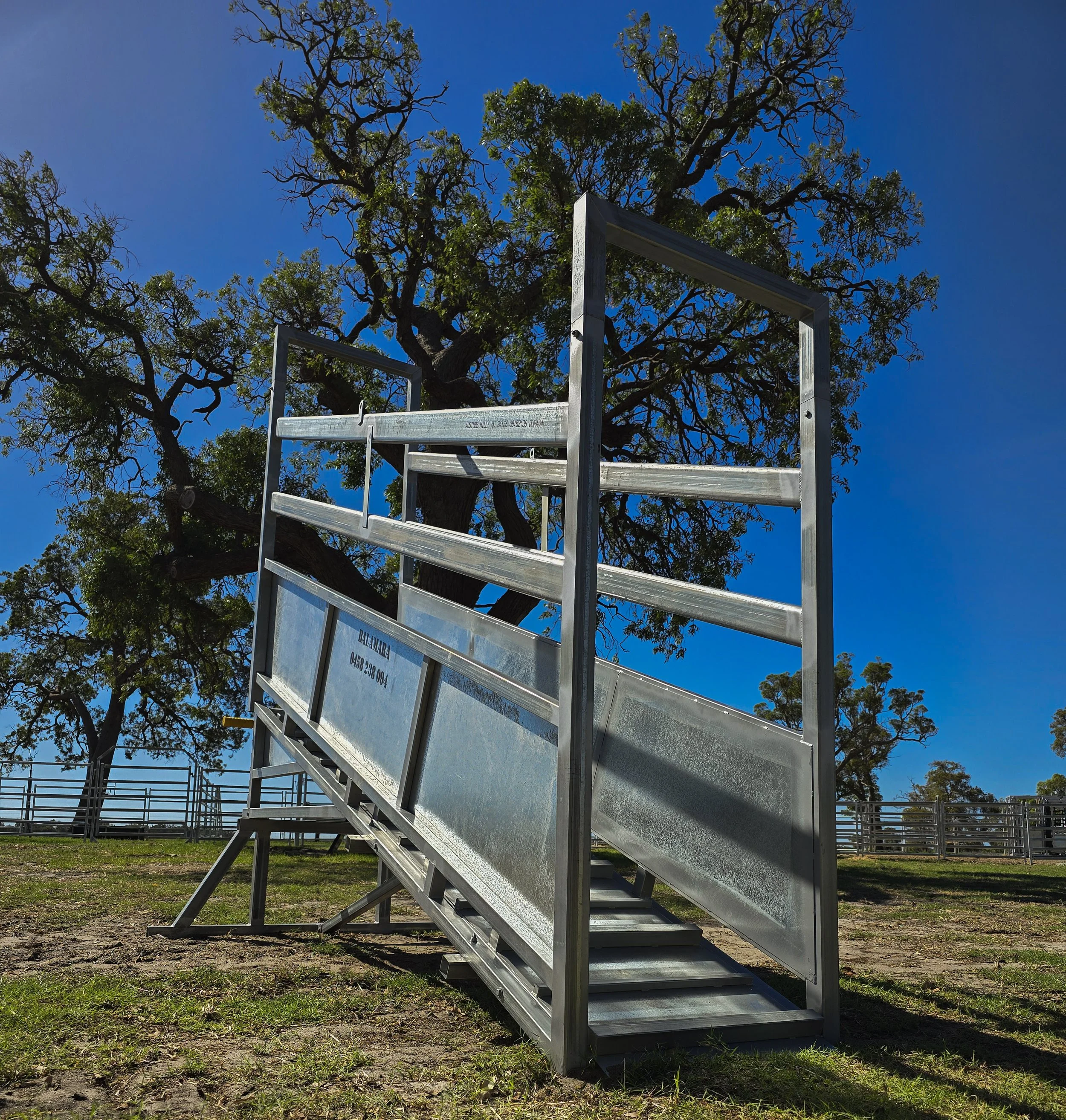 A metal hay feeder on wheels set up outdoors on grassy ground with large trees and a fence in the background under a clear blue sky.