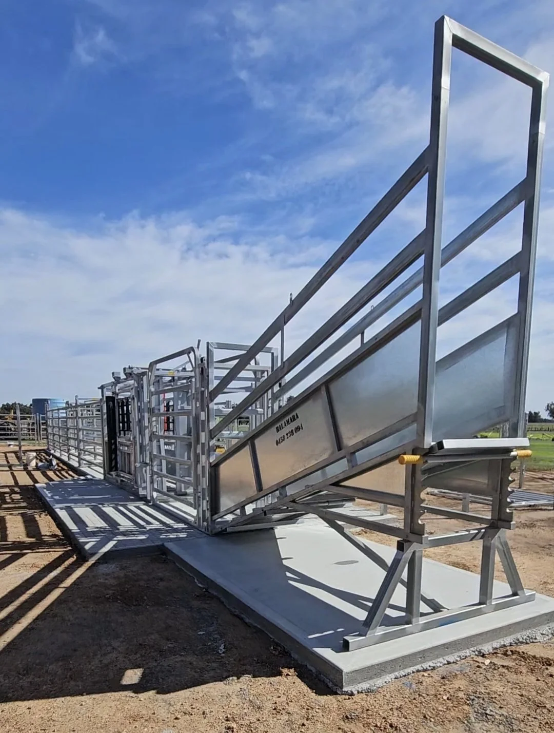 Metal livestock loading ramp on a dirt lot under a partly cloudy sky.