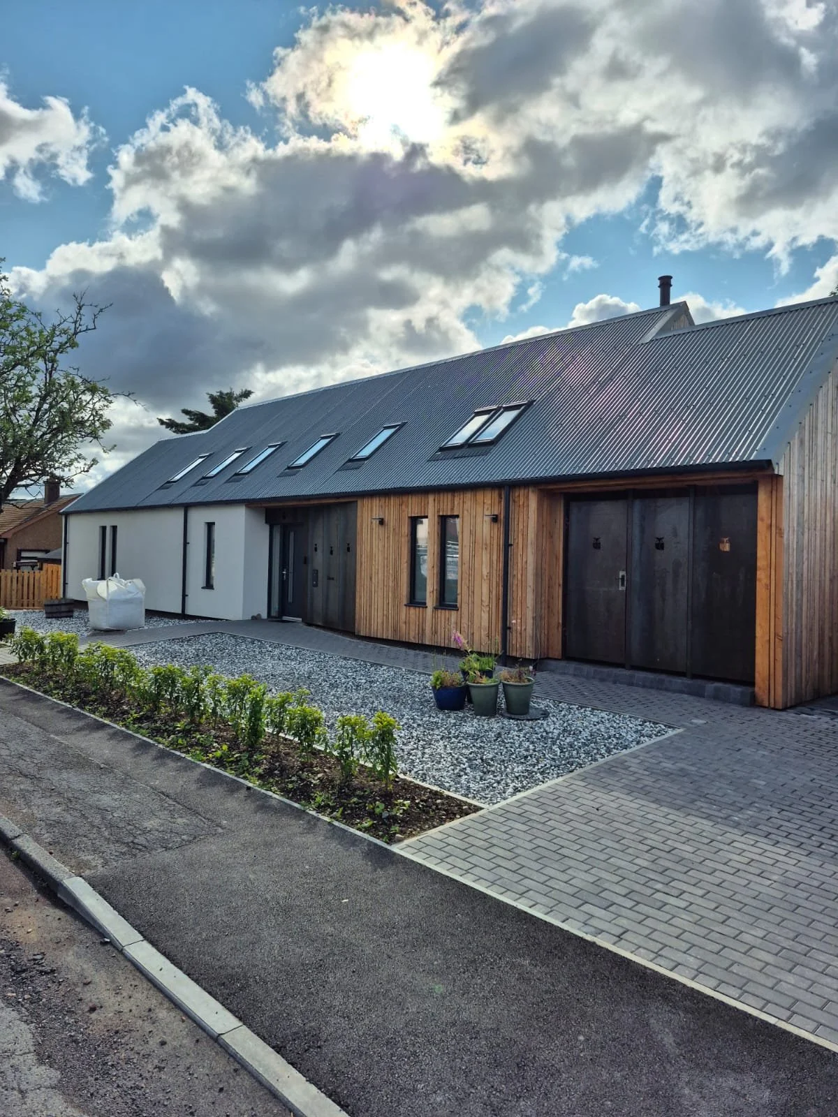 A modern residential house with a combination of white and wooden exterior walls, a sloped metal roof with skylights, and a small garden with potted plants. The house is situated on a paved and gravel driveway under a partly cloudy sky.