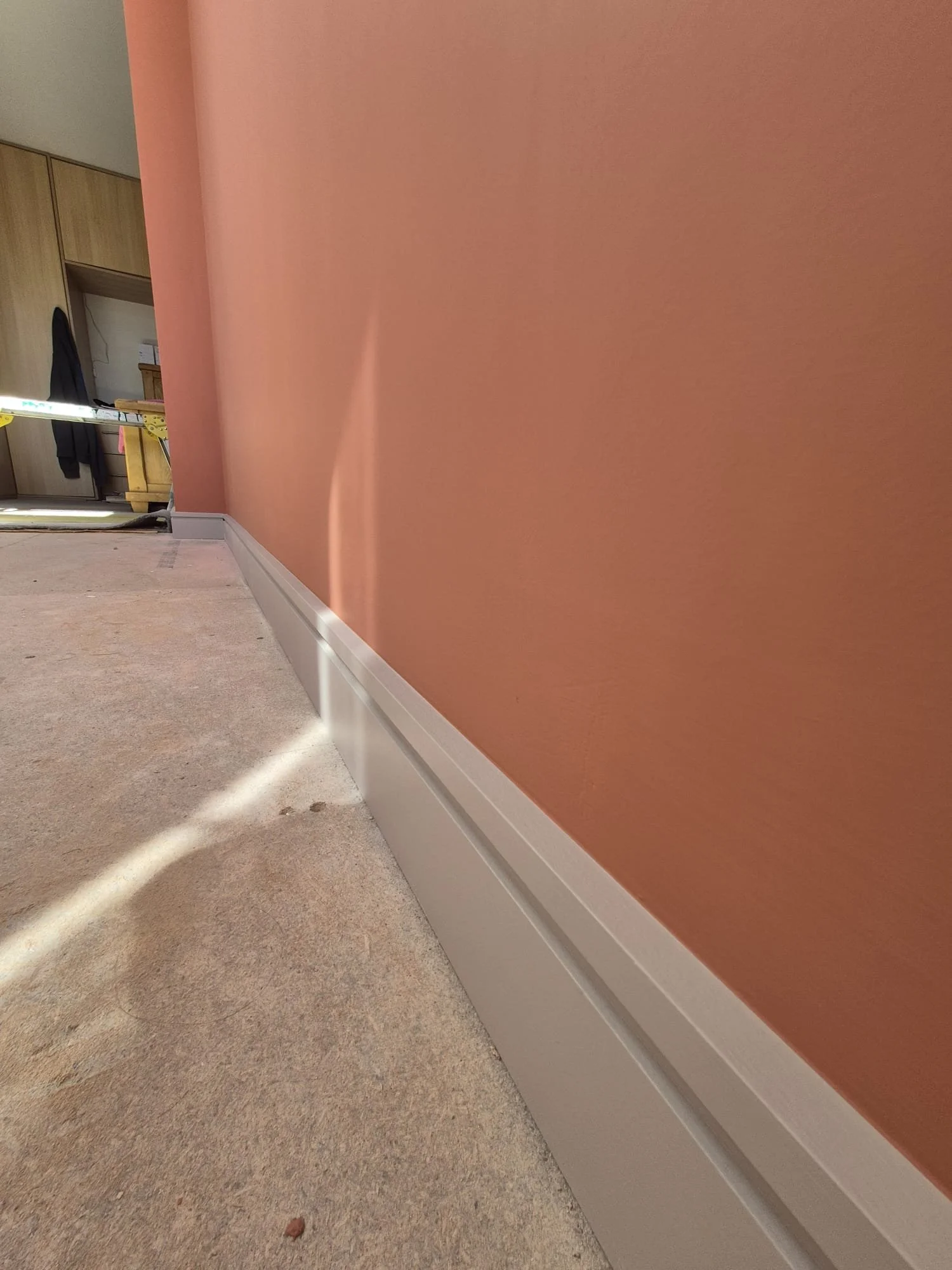 Close-up of a pink painted wall with a white baseboard, carpeted floor with some dust and marks, and a section of a room with furniture and a ladder in the background.