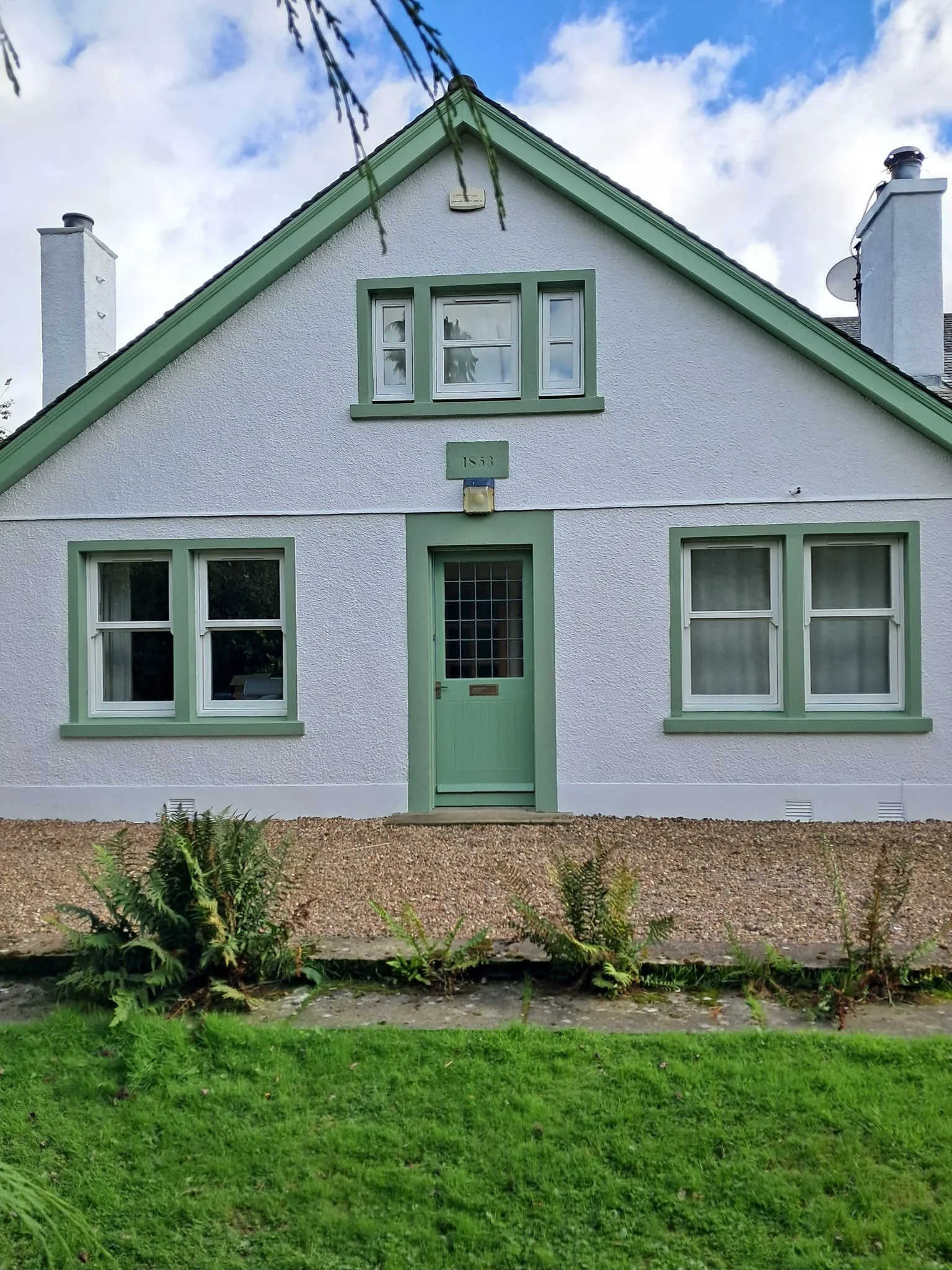 A white house with green trim, a green front door, and green window frames. The house has a triangular roof with two chimneys and a small yard with green grass and some plants in front.