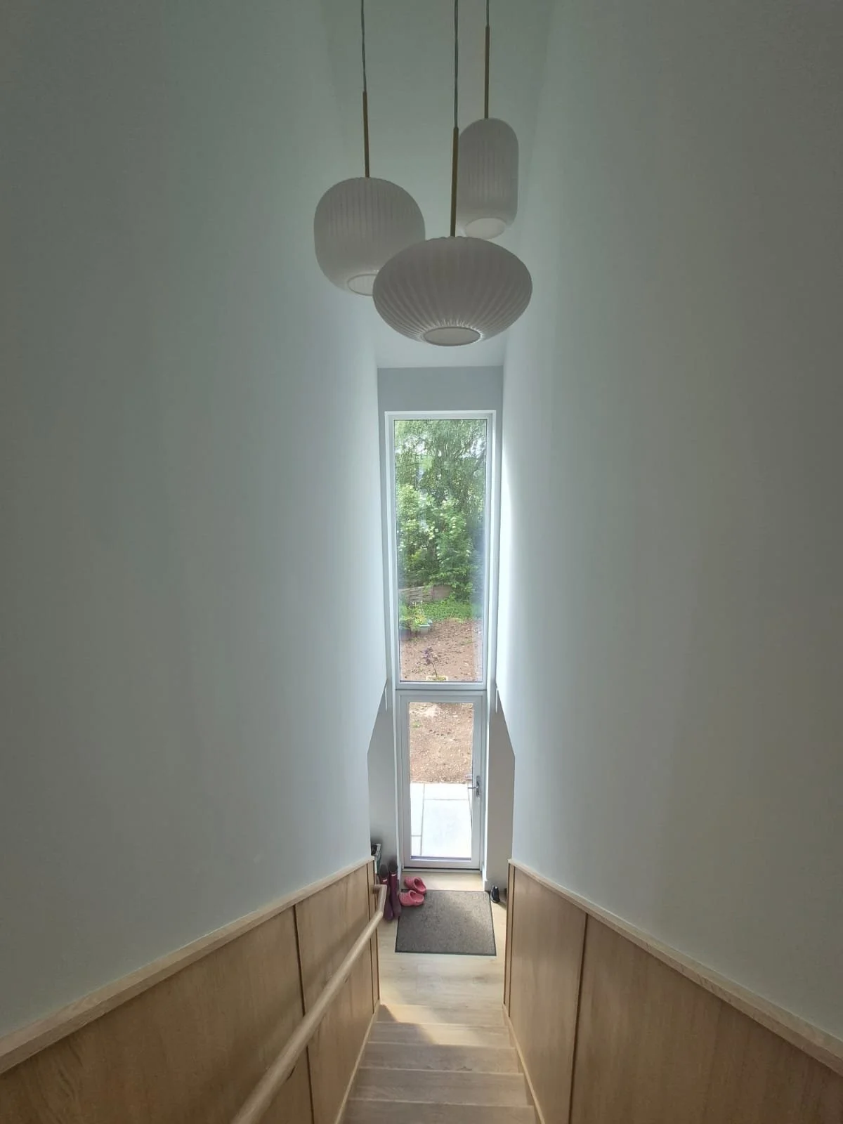 A narrow stairwell with light wood paneling, leading to a door at the bottom, with a window allowing natural light to fill the space. A ceiling light fixture with four white, ribbed, rounded shapes hangs above.