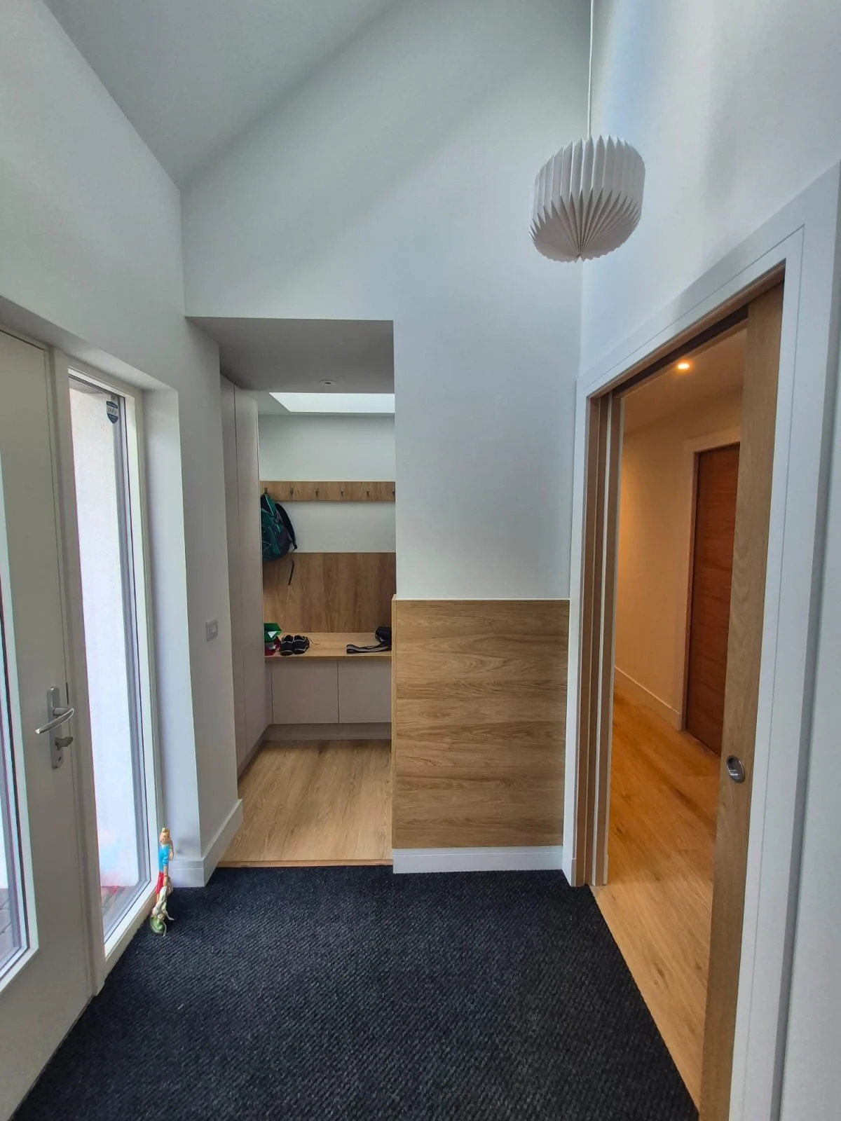 Interior entryway with white walls, a wooden accent wall, a black carpeted floor, a wooden bench with shoes, coat hooks, a nearby glass door, and a ceiling light fixture.