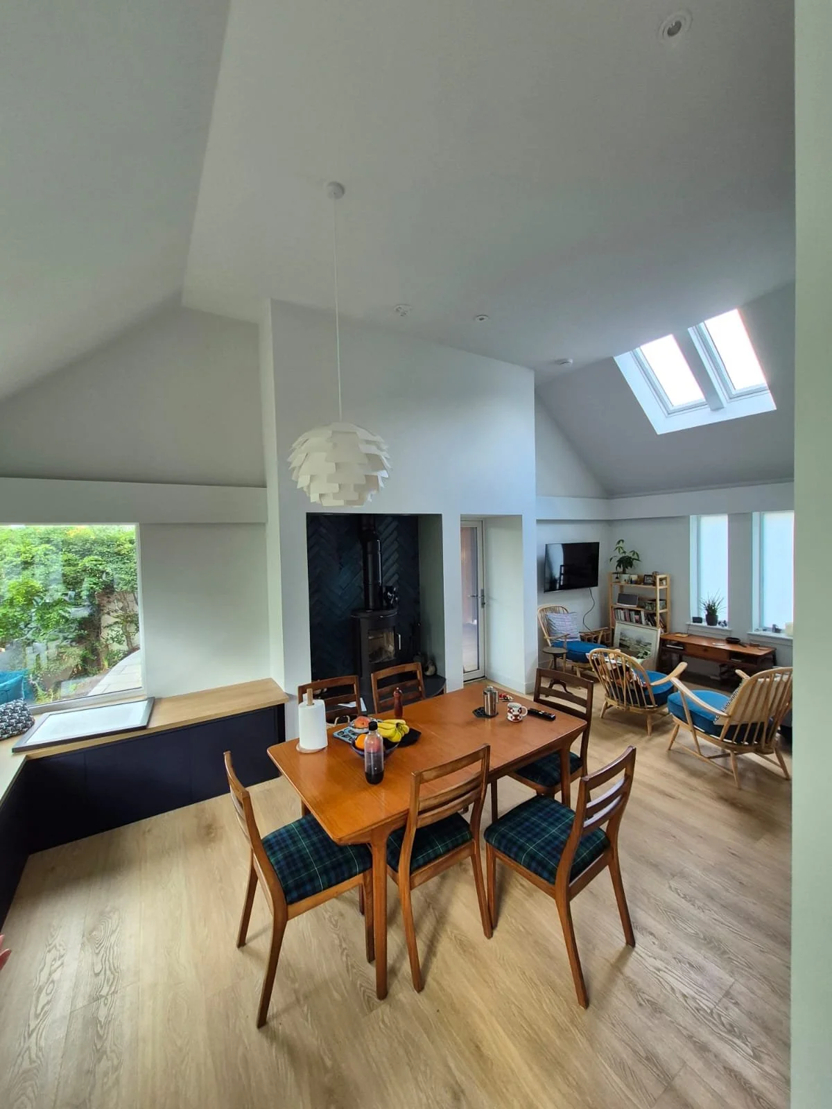 A spacious living and dining area with a wooden dining table, six chairs with plaid cushions, a black stove, and a seating area with armchairs and a TV, under a vaulted ceiling with skylights.