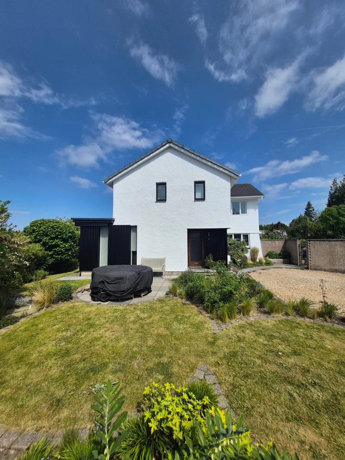 Modern white house with black accents, surrounded by a green garden and blue sky with clouds.