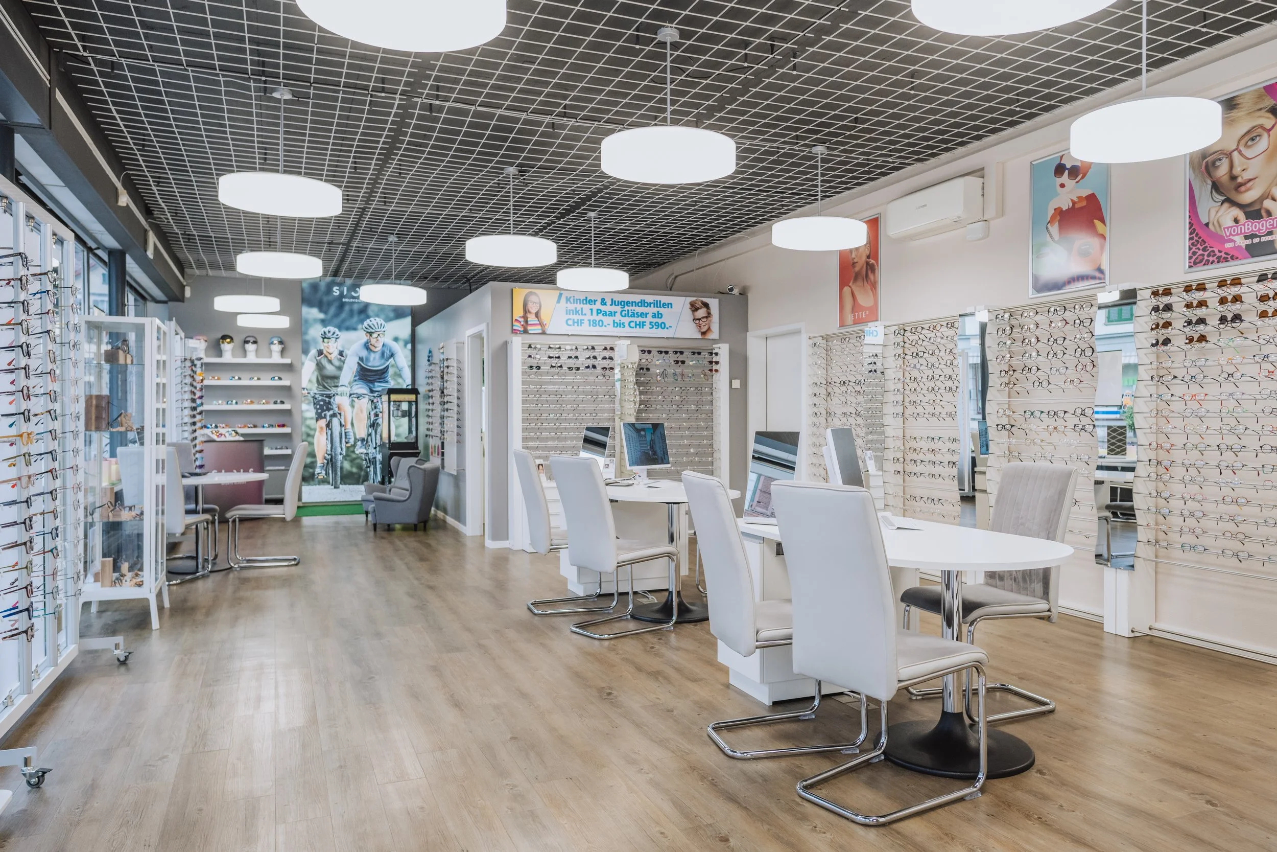 Innenansicht eines Optikergeschäfts mit Brillen auf Regalen, Computertischen und Sitzmöglichkeiten, Wanddekorationen und digitaler Preisschilder.