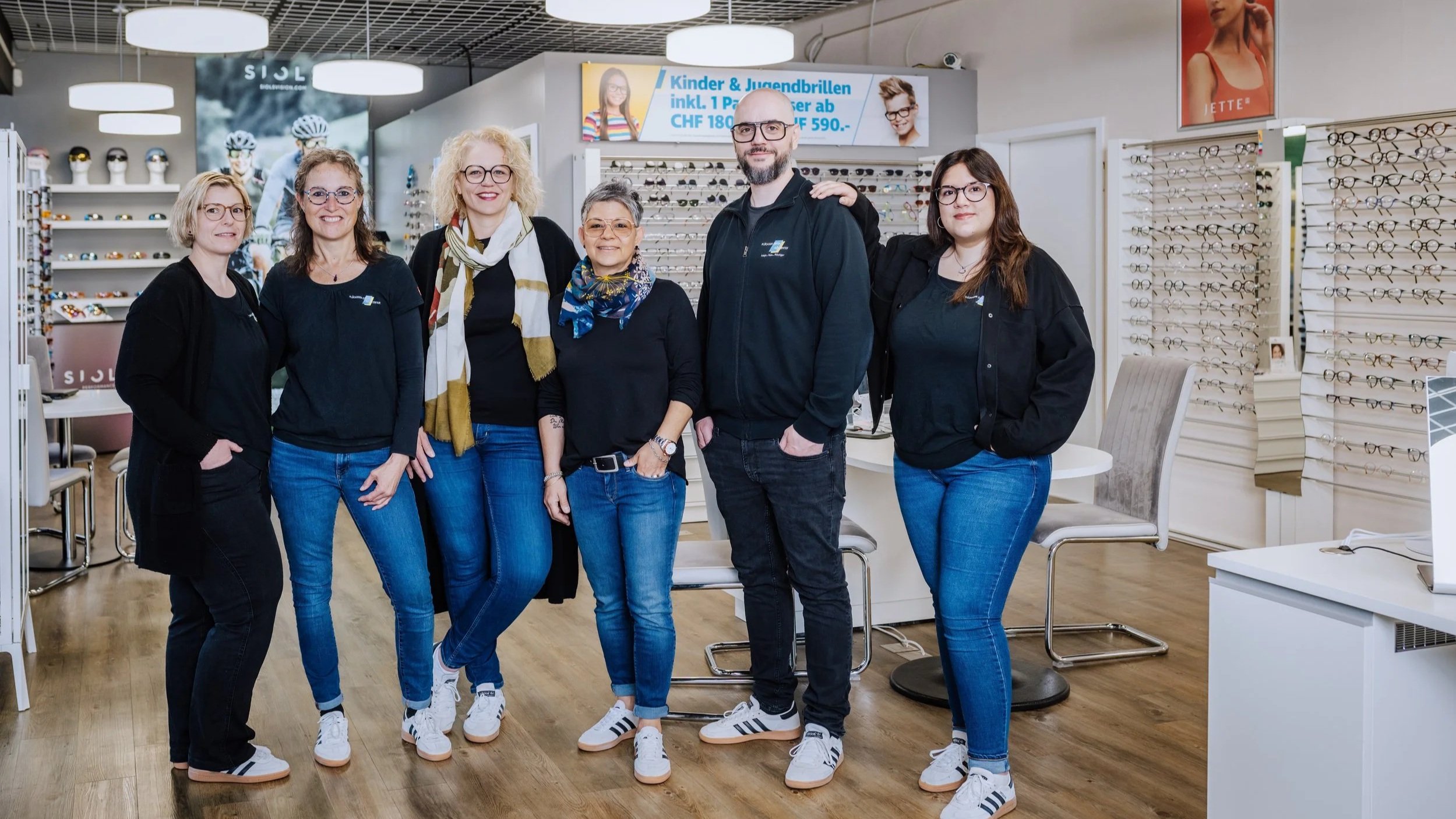 Sechs Menschen stehen in einem Optikergeschäft, umgeben von Brillenfassungen und Sonnenbrillen, alle lächelnd und in legerer Kleidung. Im Hintergrund sind Regale mit Brillen ausgestellt, und eine digitale Anzeigetafel mit Angeboten ist sichtbar.