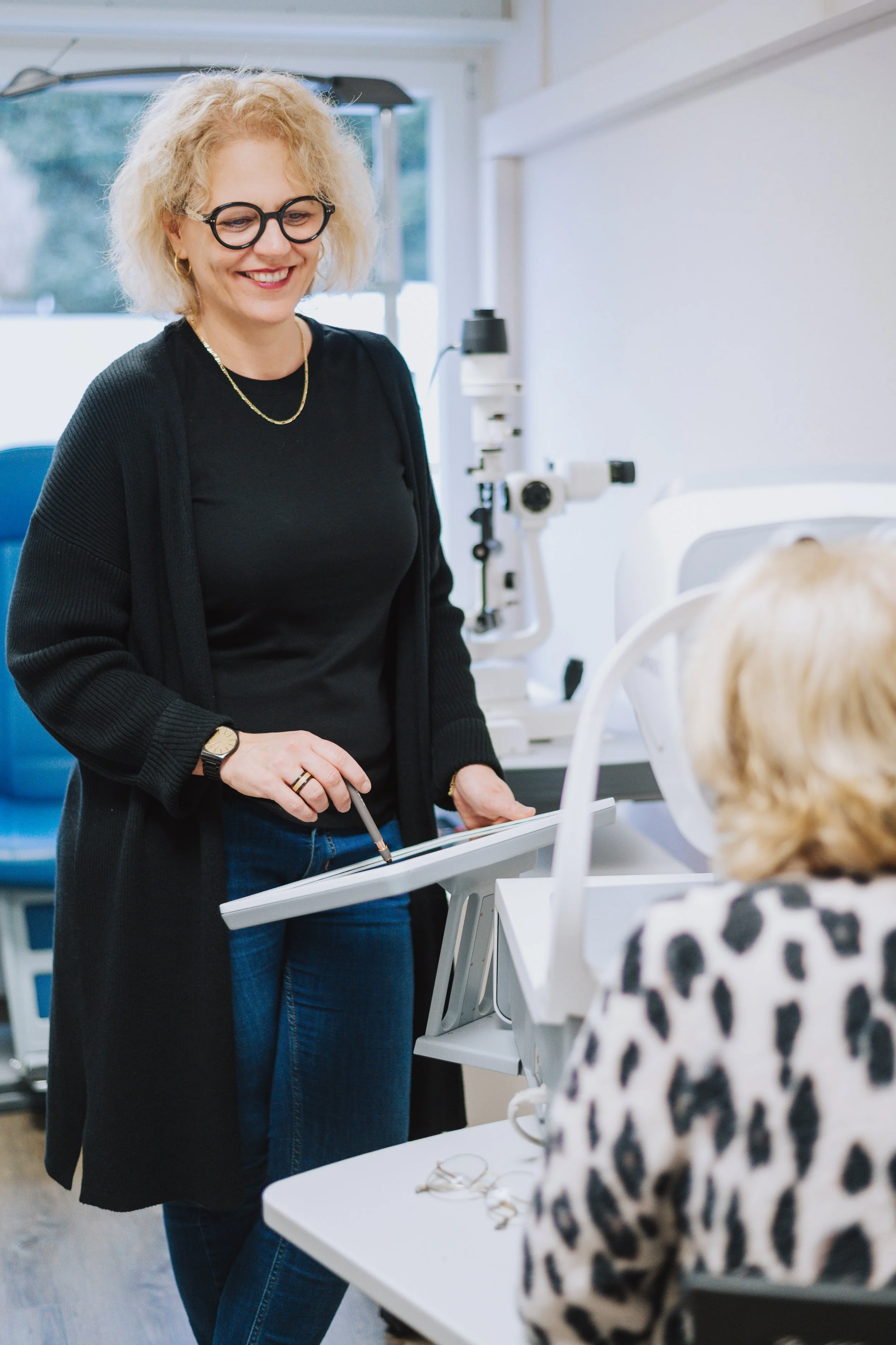 Eine lachende Frau mit Brille, schwarze Kleidung und einem Notizbuch steht in einem medizinischen Raum vor einem Patienten, der im Rollstuhl sitzt, mit einem medizinischen Gerät im Hintergrund.