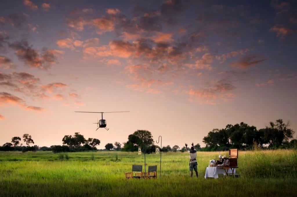 A helicopter flying over a grassy field with a person taking photos, and a table with items on it, during sunset with a colorful sky and scattered clouds on an African tour.
