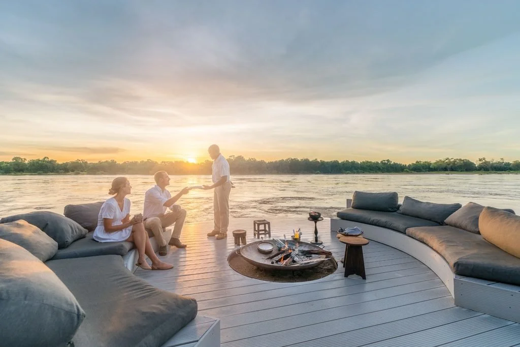 Guests enjoying sundowners by a firepit on the river deck at Chinzombo, South Luangwa National Park, Zambia