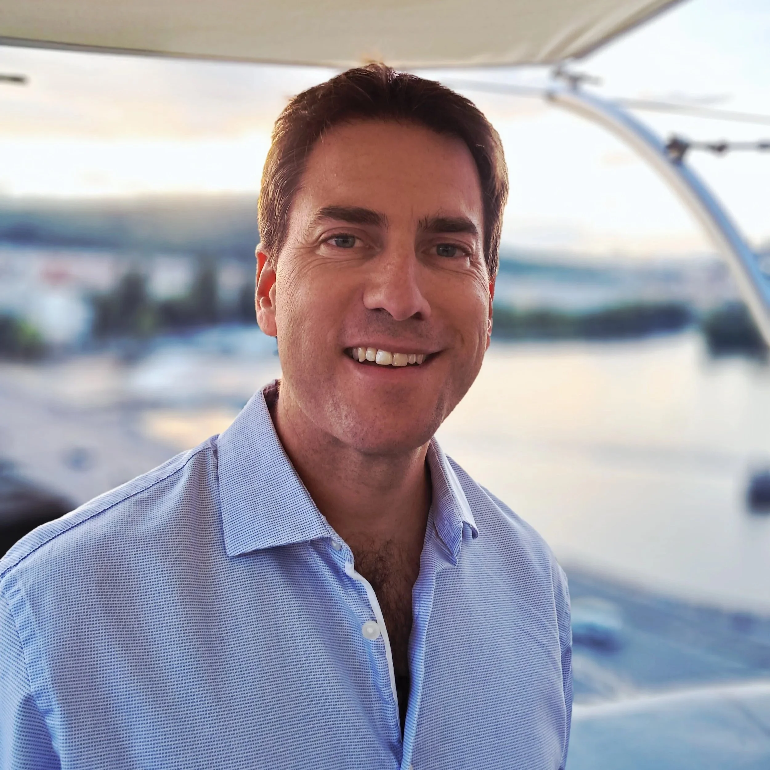 A smiling man with short brown hair wearing a light blue button-up shirt, standing outdoors near water with boats in the background.