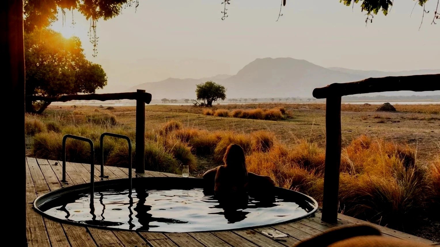 A person relaxing in an outdoor hot tub on a wooden deck, overlooking a vast savannah landscape with scattered trees, distant mountains, and a setting sun.
