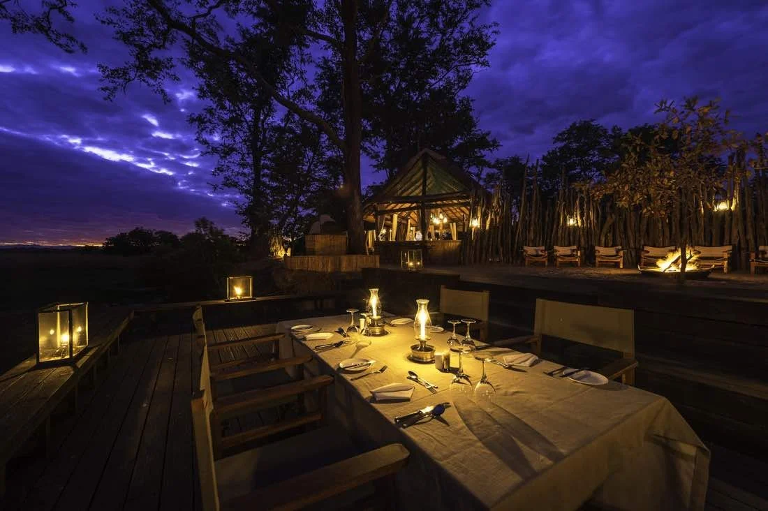 Aerial view of a river at sunset with a small, illuminated outdoor dining area on a platform near the riverbank, surrounded by trees and bushes.