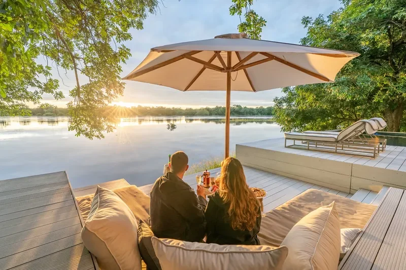 Couple enjoying sunrise drinks on the riverfront deck at Time + Tide Chinzombo under a parasol, South Luangwa, Zambia