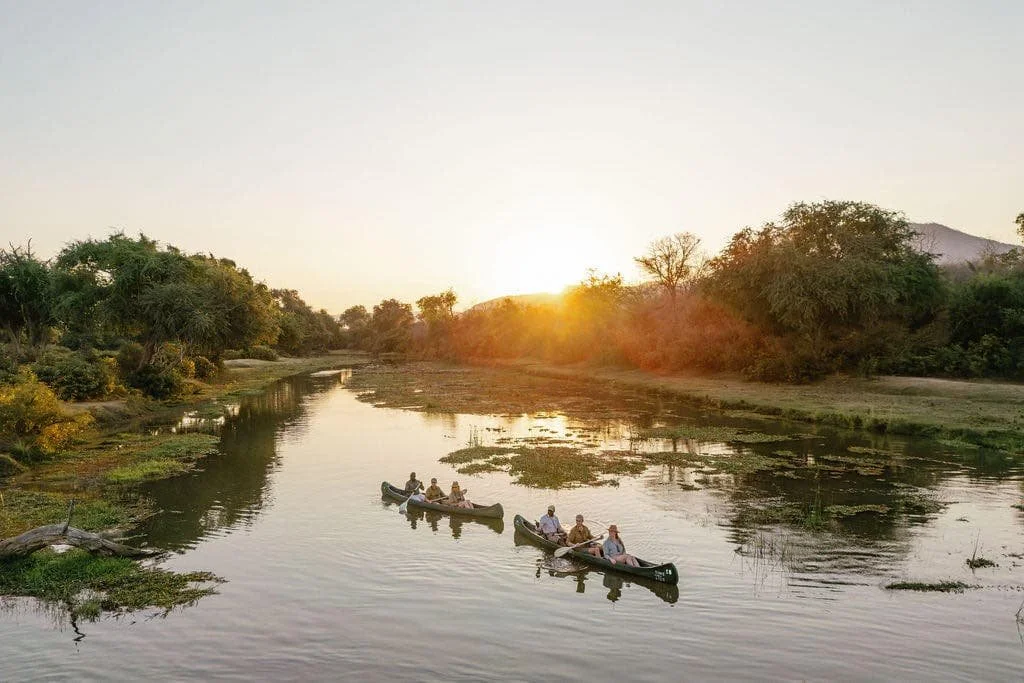 Five people in two canoes on a calm river during sunset, surrounded by lush greenery.