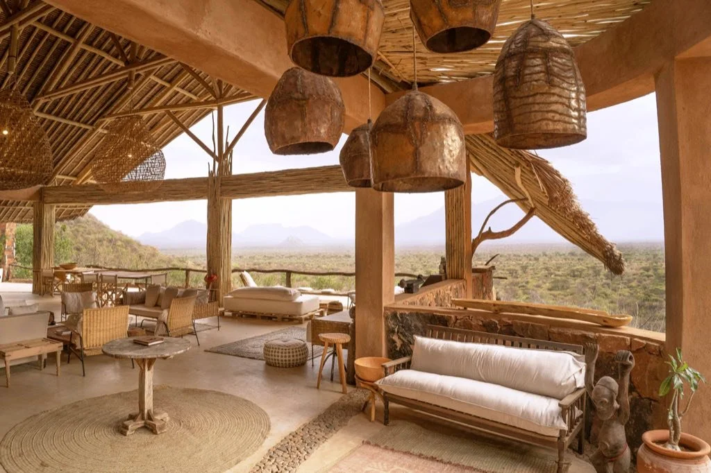 Open-air rustic lounge area with wooden furniture, hanging pendant lamps, and a panoramic view of a desert landscape with mountains in the distance.