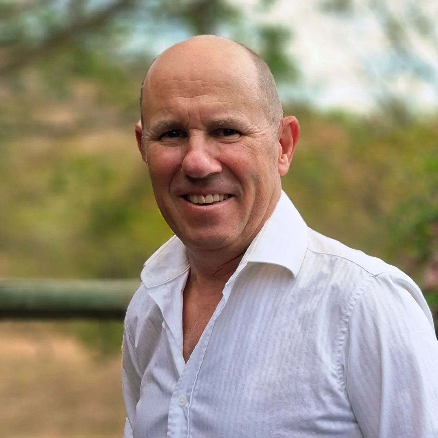 A middle-aged man with a shaved head wearing a white button-up shirt, standing outdoors with a blurred background of trees and greenery.