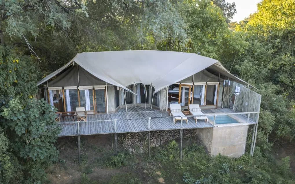 Open-air dining area with a long wooden table set with black plates, glasses, and silverware under a thatched roof, overlooking a river with trees in the background.