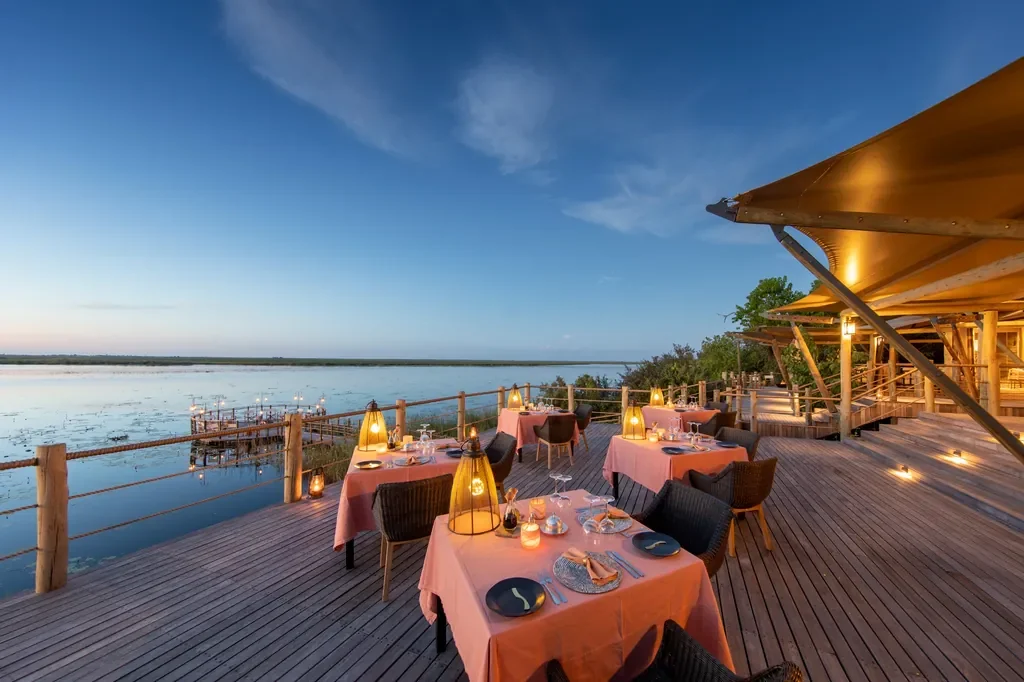 Lagoon-side dining deck at DumaTau overlooking the Linyanti, lantern-lit tables at blue hour.