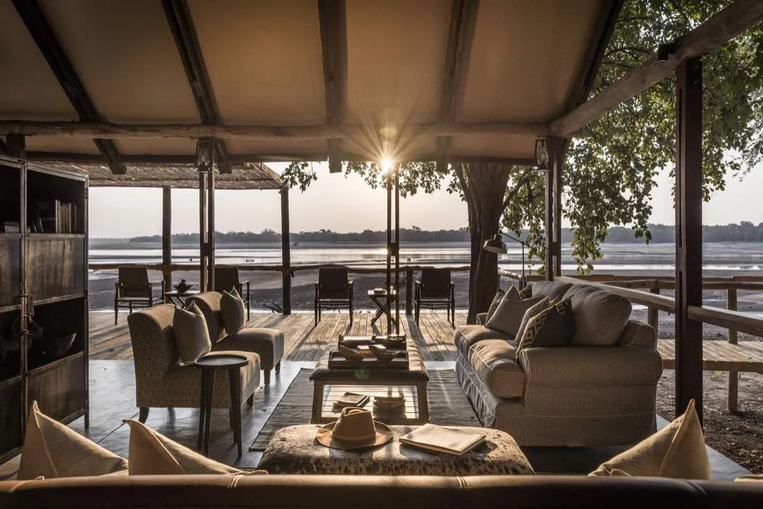 Open-air rustic lounge area with wooden furniture, hanging pendant lamps, and a panoramic view of a desert landscape with mountains in the distance.