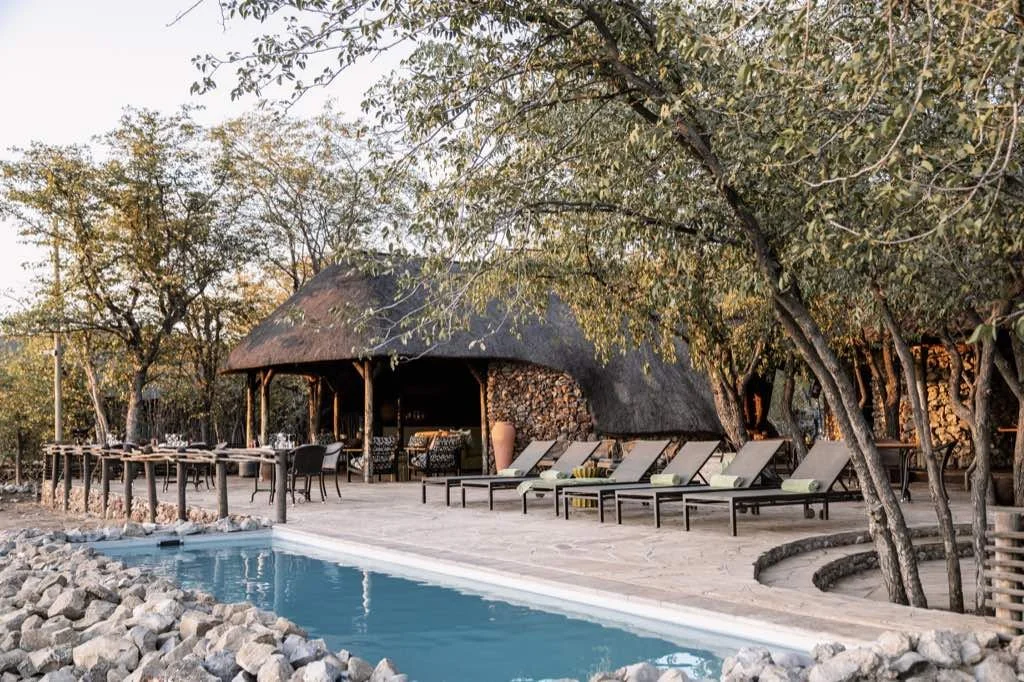 Poolside lounge chairs and a thatched-roof cabana surrounded by trees at sunset.