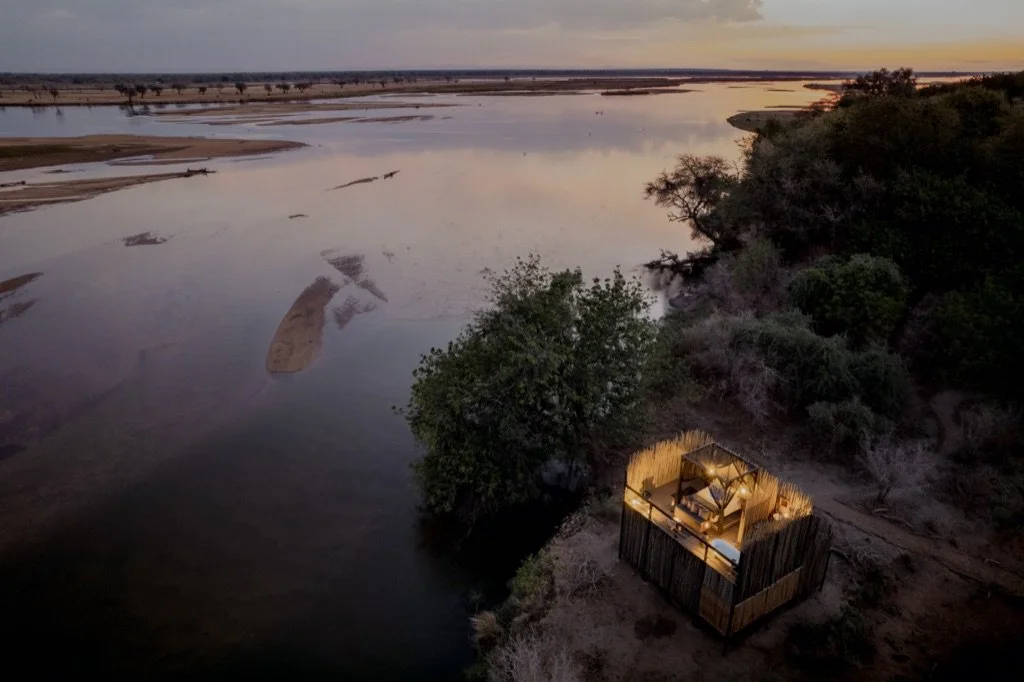 Aerial view of a river at sunset with a small, illuminated outdoor dining area on a platform near the riverbank, surrounded by trees and bushes.