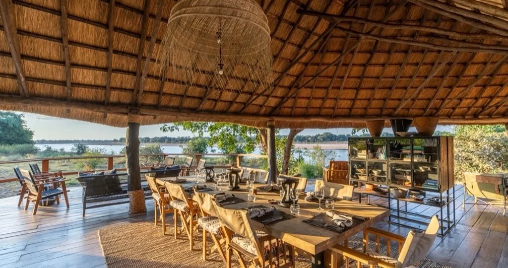 Open-air dining area with a long wooden table set with black plates, glasses, and silverware under a thatched roof, overlooking a river with trees in the background.
