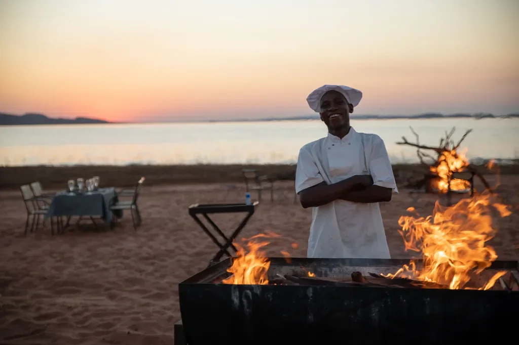 Lakeside beach braai with smiling chef at sunset, Bumi Hills Safari Lodge, Lake Kariba, Zimbabwe