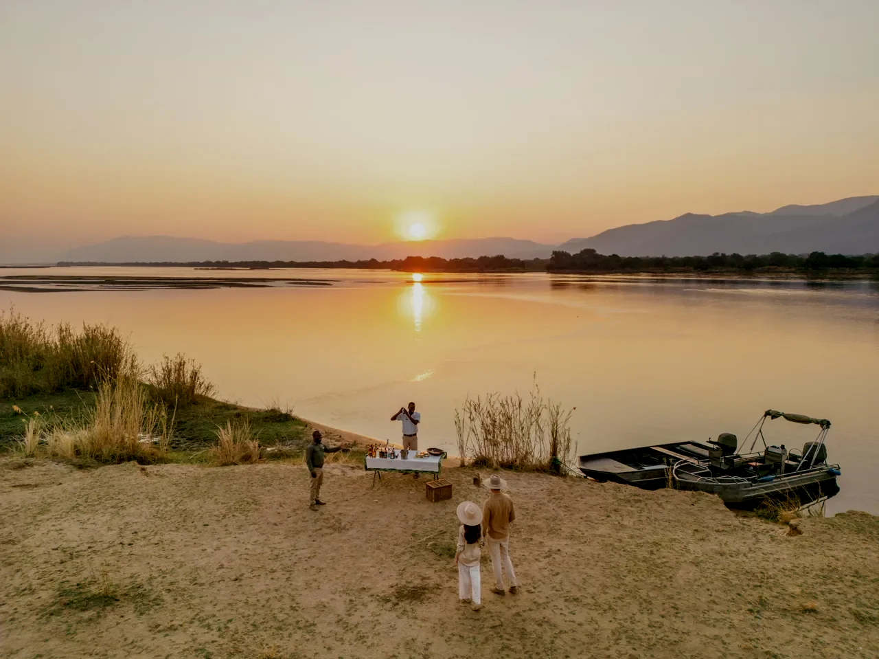People enjoying a gathering by the river at sunset with a boat docked nearby and a table of drinks.