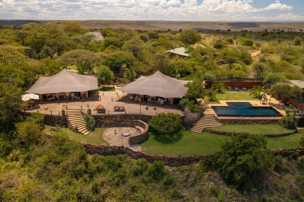 Aerial view of a luxurious outdoor retreat with two large beige tents, a swimming pool, lush greenery, and stone pathways in a rural setting.