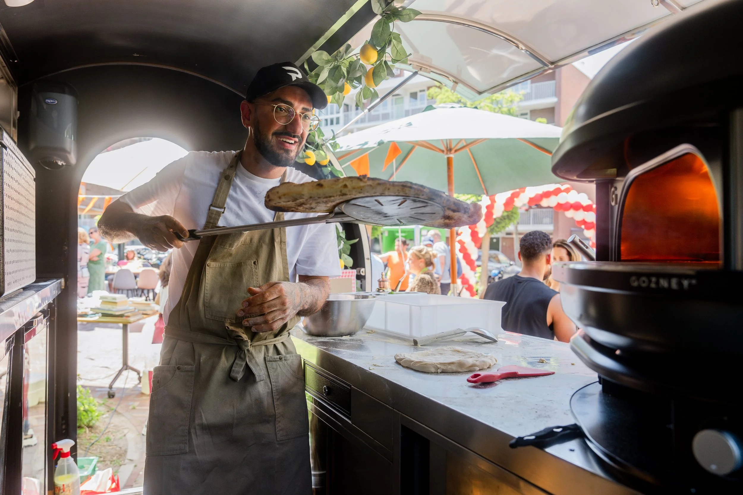 A smiling man wearing glasses, a cap, and a beige apron is holding a pizza peel with a pizza and preparing to place it into a pizza oven. He is standing inside a food truck during a lively outdoor event with people, umbrellas, and decorations visible in the background.