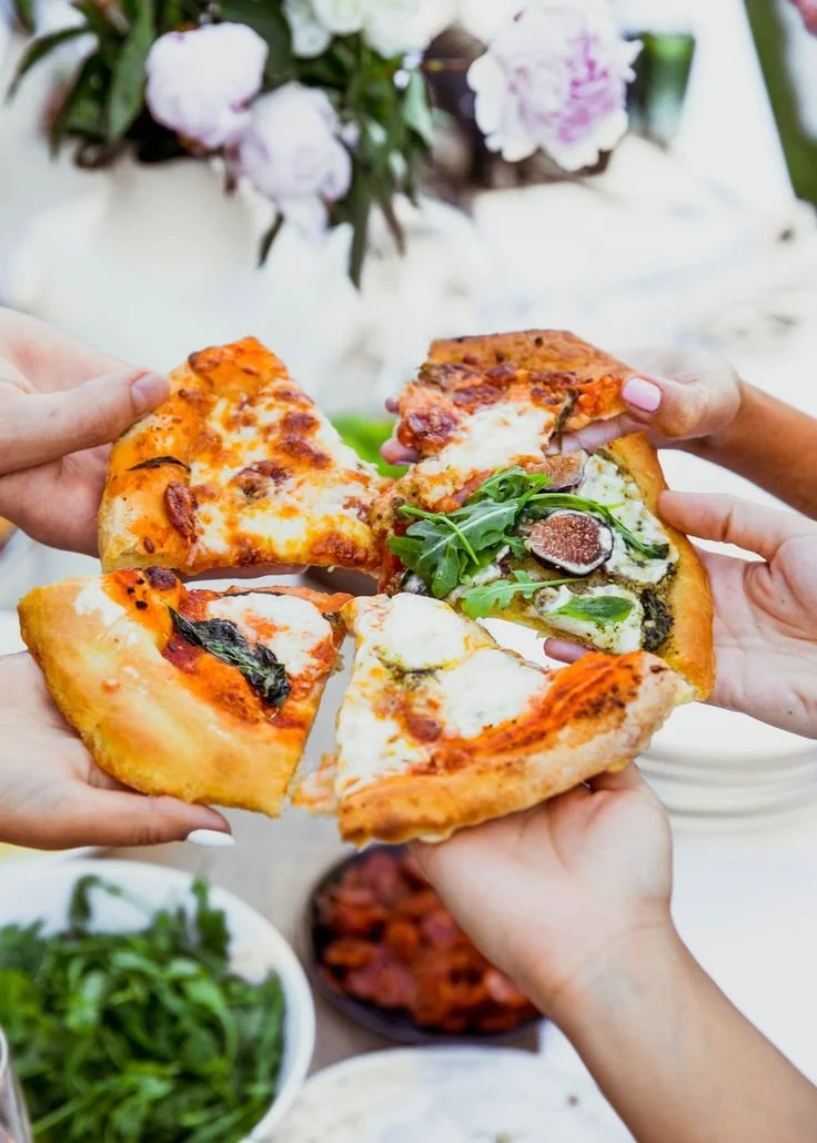 Group of hands holding and sharing a pizza with various toppings, with a blurred floral arrangement in the background.