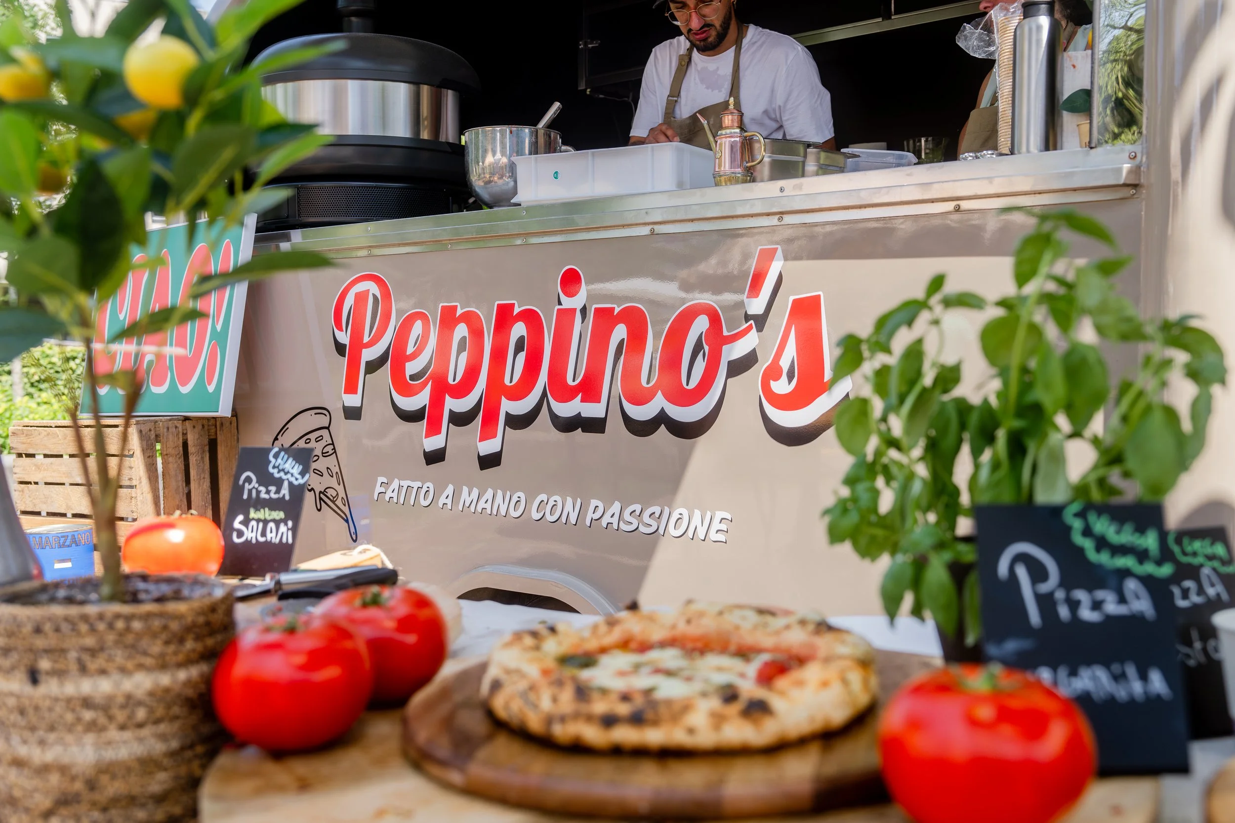 A food truck named 'Peppino's' with a sign that says 'Fatto a mano con passione'. In the foreground, there is a pizza on a wooden cutting board, tomatoes, and potted herbs, with a person preparing food inside the truck.