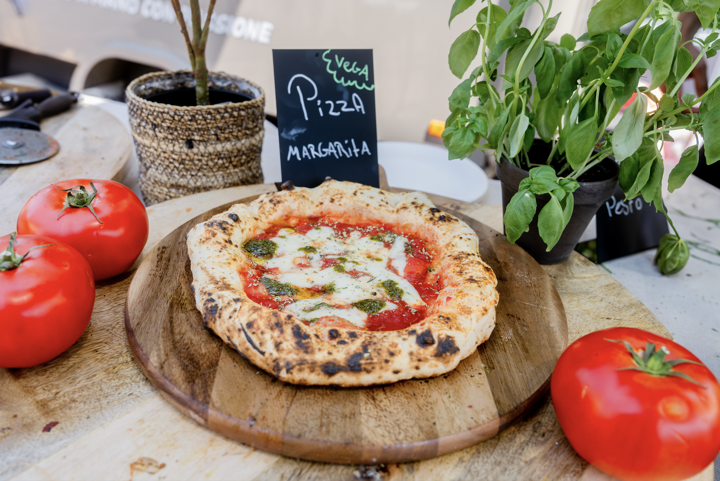 Wood-fired pizza with tomato sauce, cheese, and basil, placed on a round wooden board, with fresh tomatoes and potted basil plant on a table.