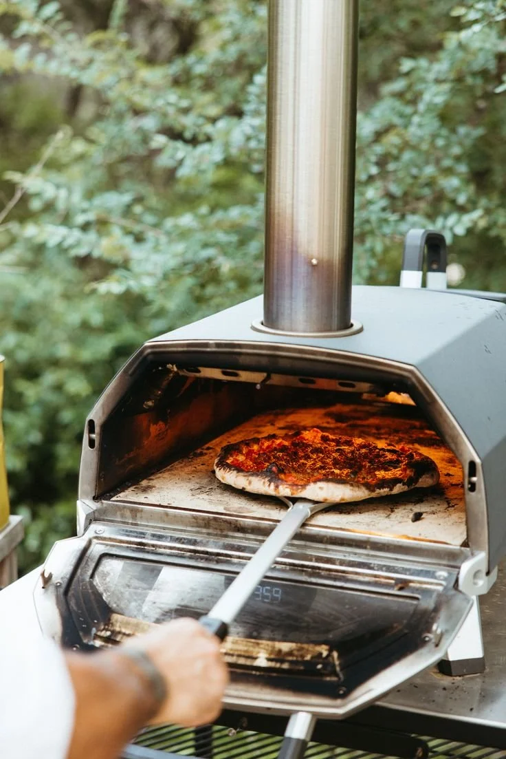A person is taking a freshly cooked pizza out of a pizza oven outdoors, surrounded by greenery.