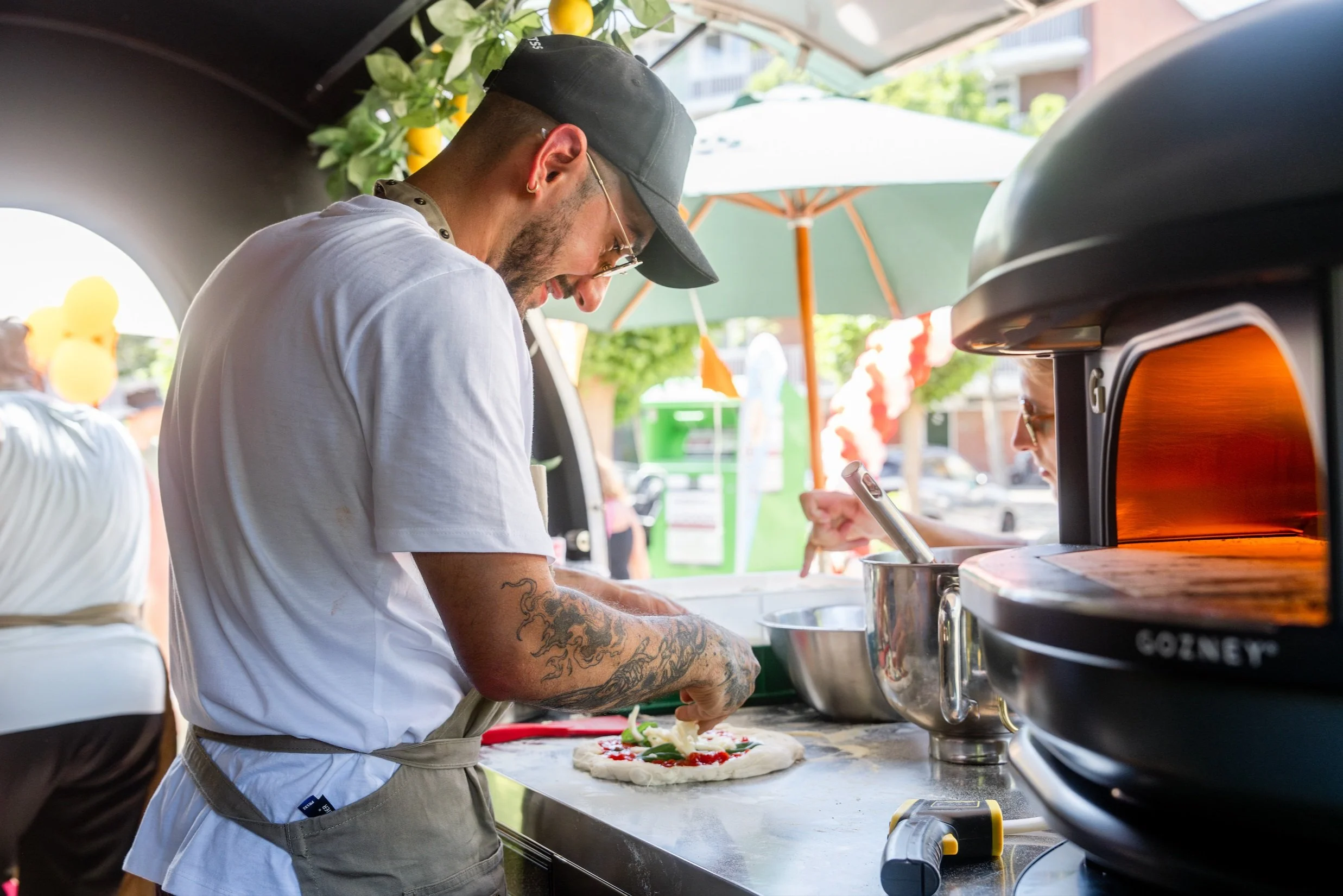 A man with a tattooed arm making a pizza at an outdoor pizza stand. He is wearing a black cap, glasses, a white t-shirt, and an apron. There are toppings on the pizza dough and a pizza oven nearby.
