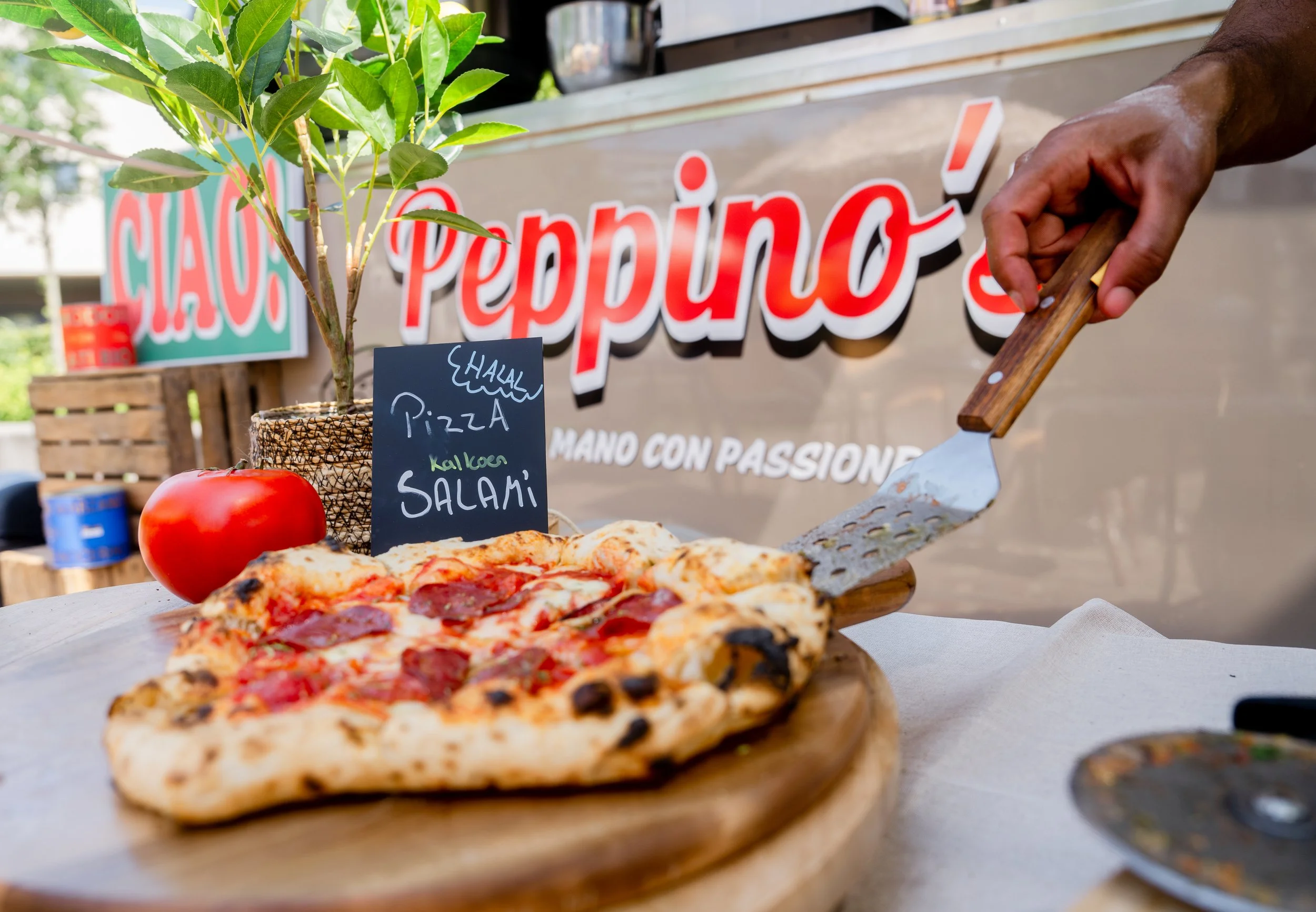 Fresh pizza with pepperoni on a wooden pizza peel, with a red tomato and a potted plant nearby, set on a table outside a pizza stand with a sign reading 'Peppino's' in the background.