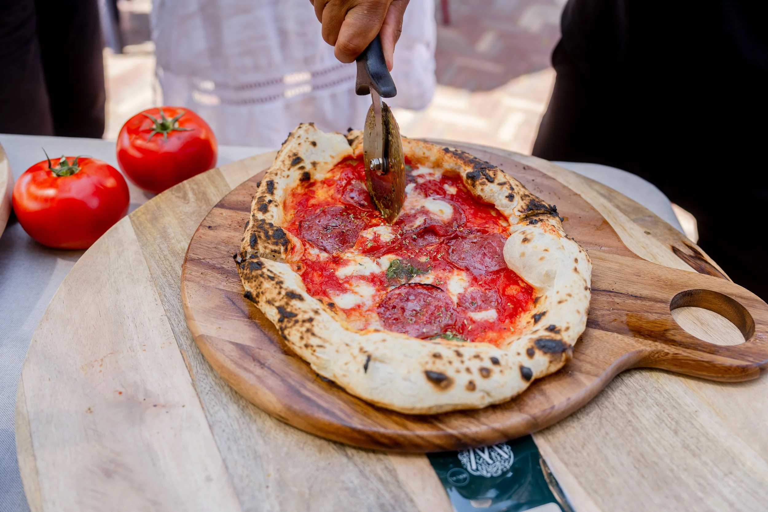 Person slicing a Margherita pizza on a wooden serving board, with fresh tomatoes nearby.