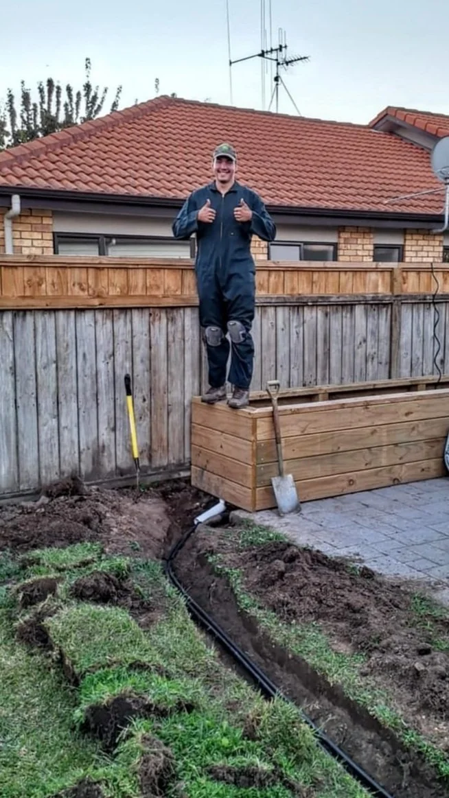 Man standing on a wooden planter box giving two thumbs up, with a garden trench and a black irrigation pipe in the yard.