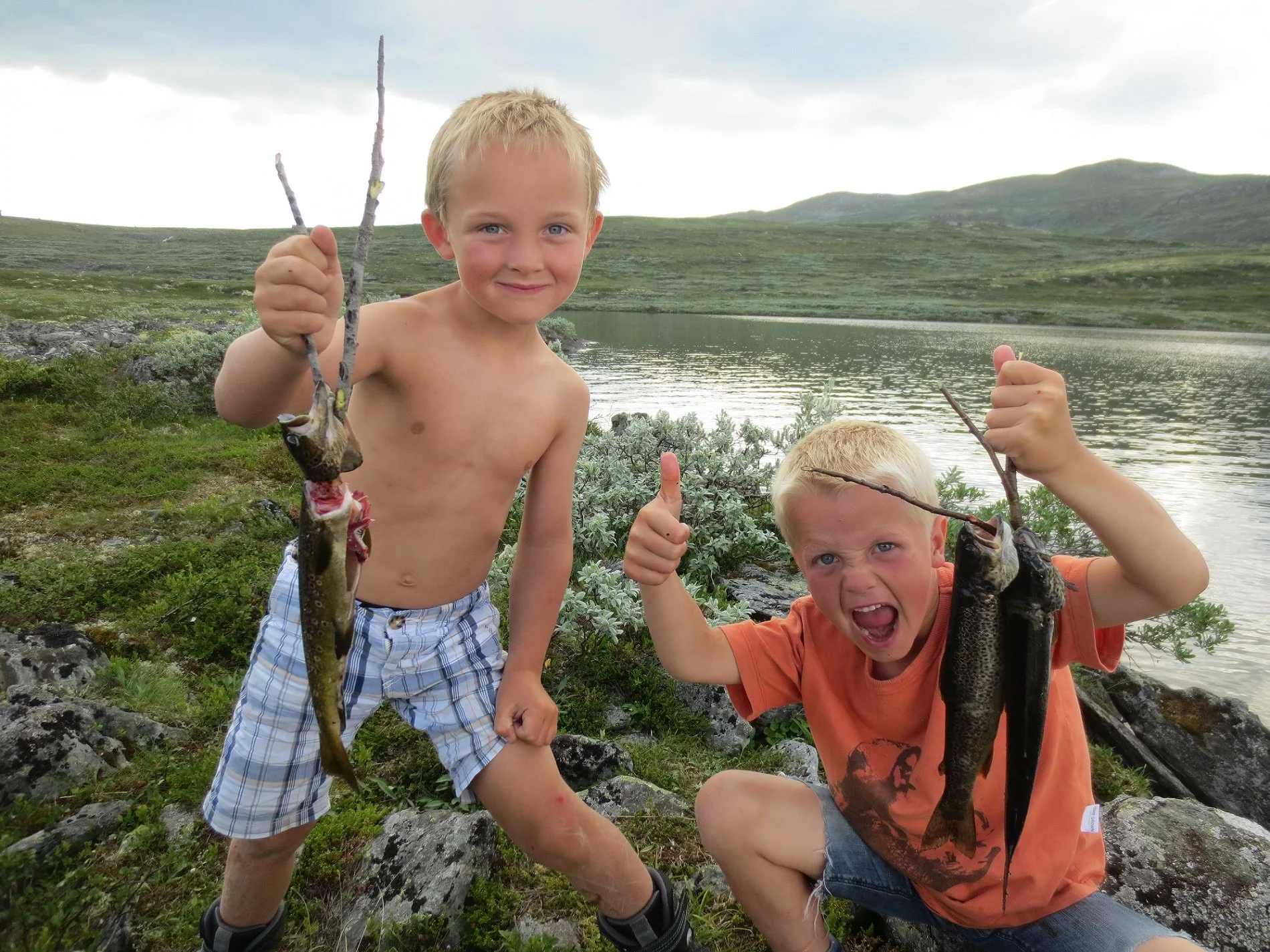 To unge gutter legger opp i naturen ved en innsjø, holder opp fisk de har fanget, smiler og viser stolt