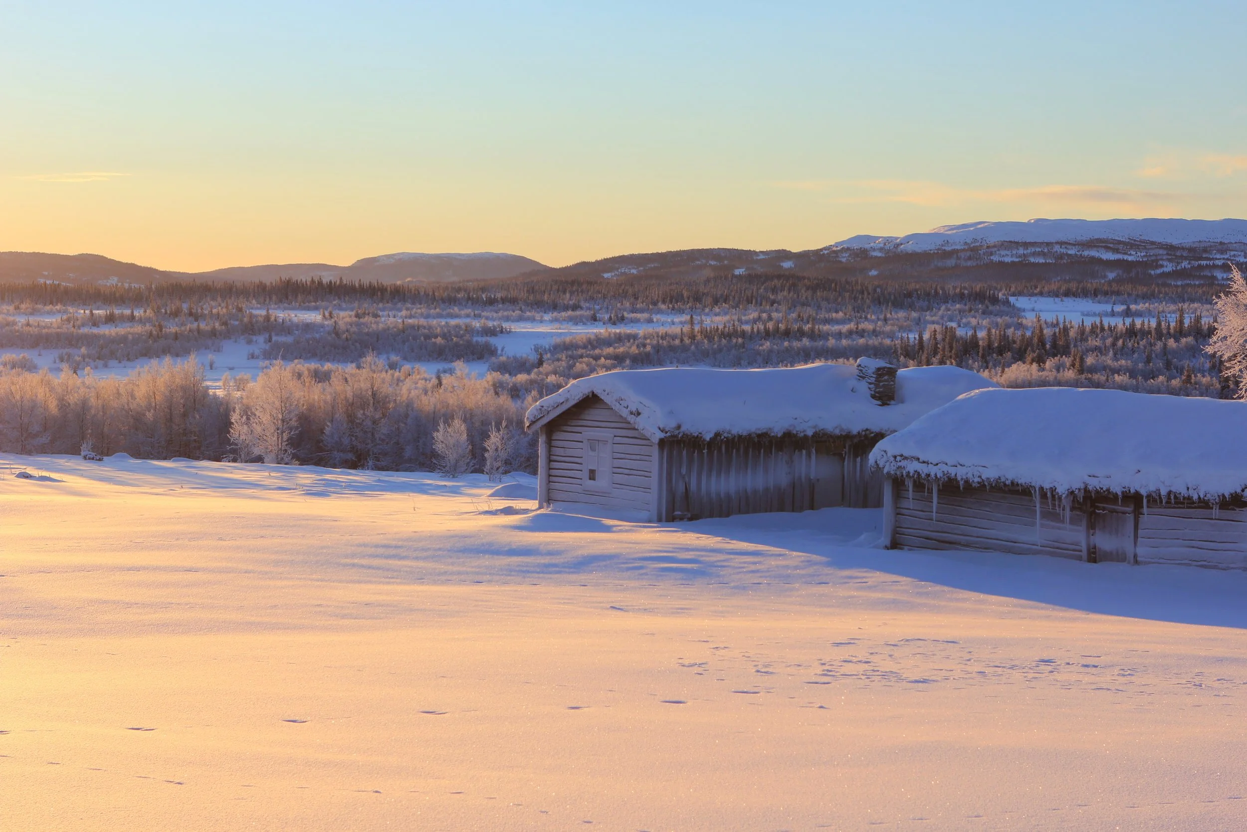 Snødekt landskap med to små trehus og en barne- eller dyresporspor i snøen, med fjell og skog i bakgrunnen ved solnedgang.