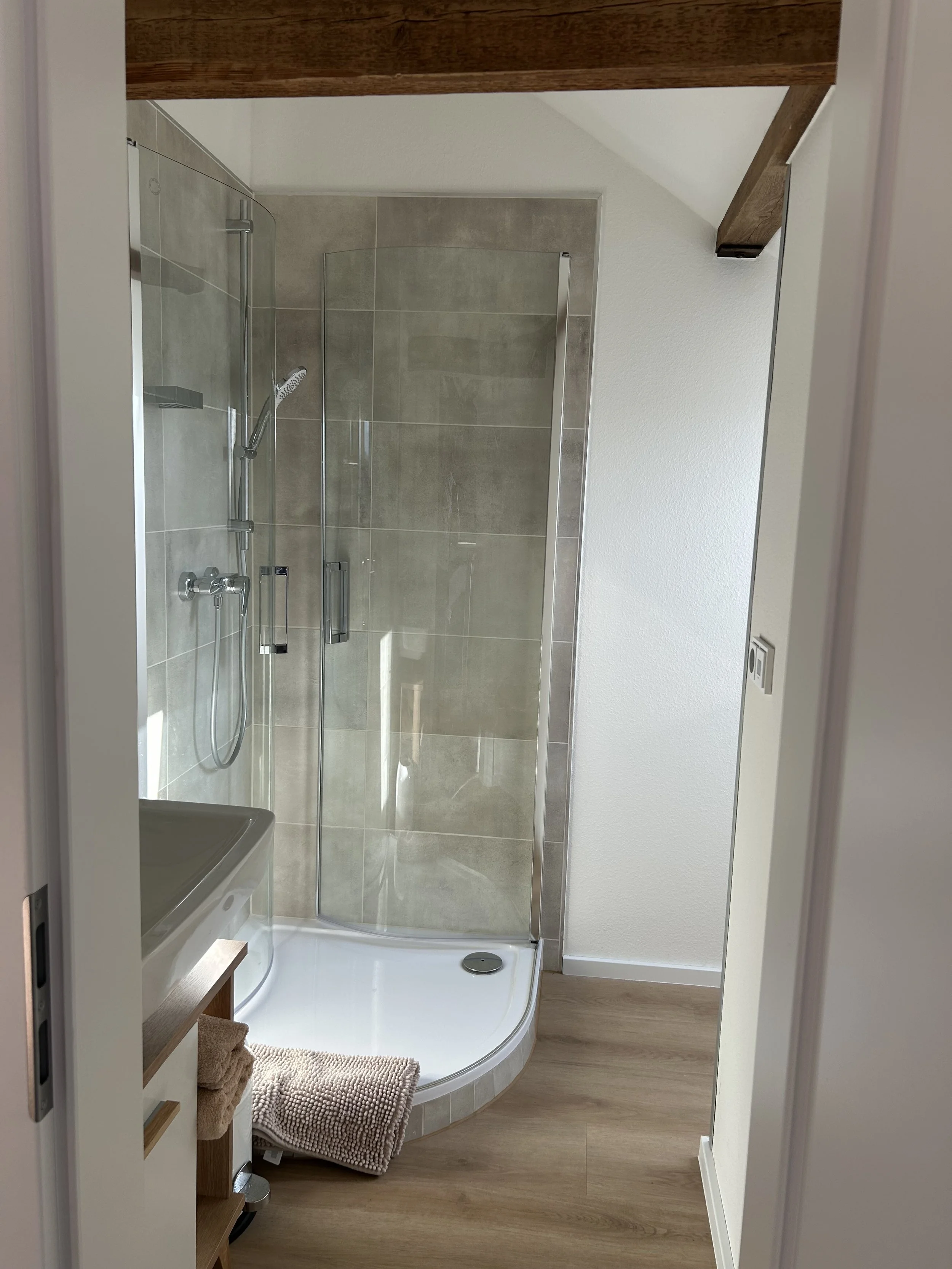 Kleines Badezimmer mit separater Dusche, Holzboden, weiße Wand, Holzdecke, Fach mit Handtüchern und Waschtisch.