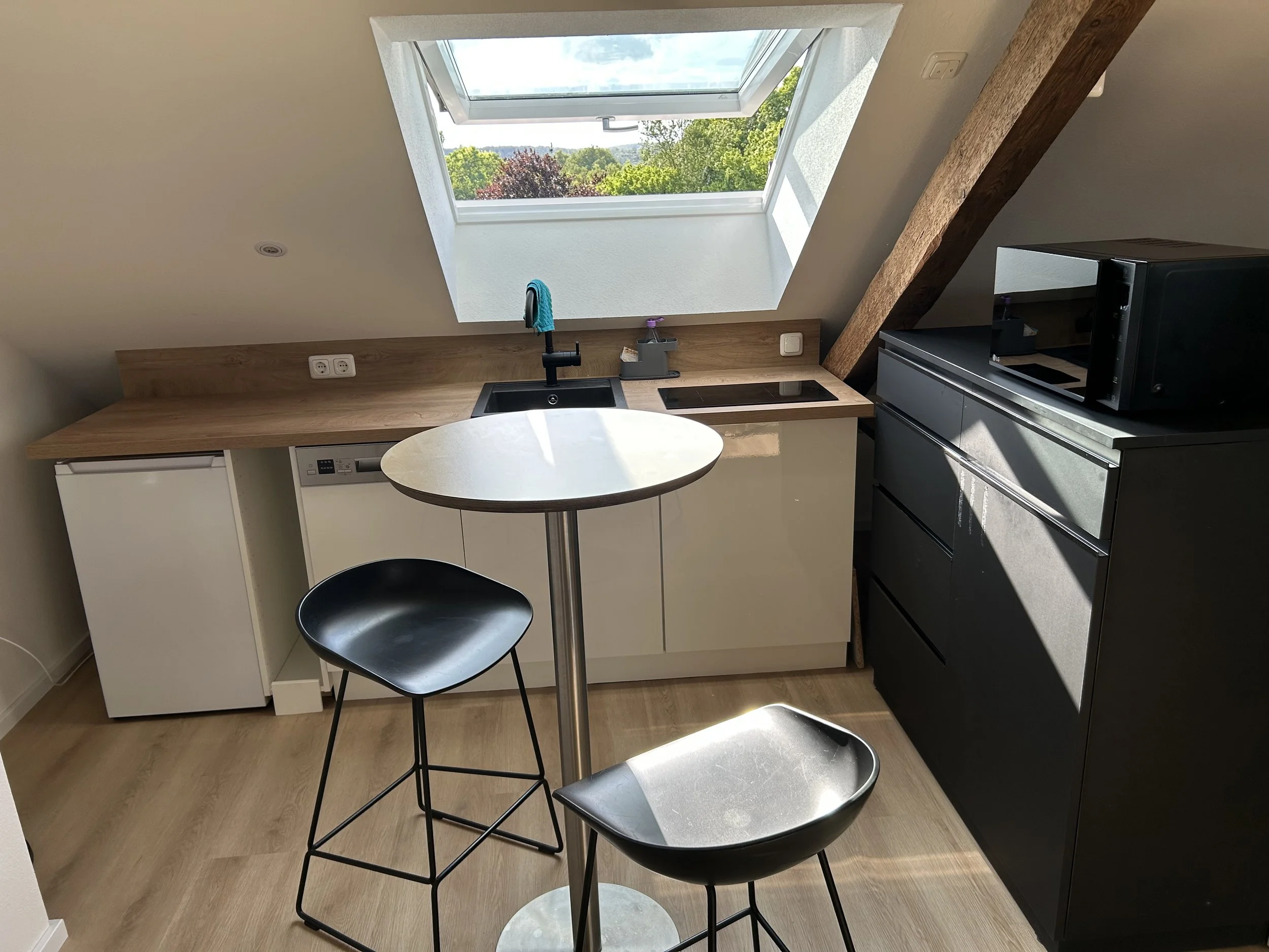 Küchenzeile mit Spüle, Kochfeld, Mikrowelle, Kühlschrank und Barhockern unter einem Dachfenster