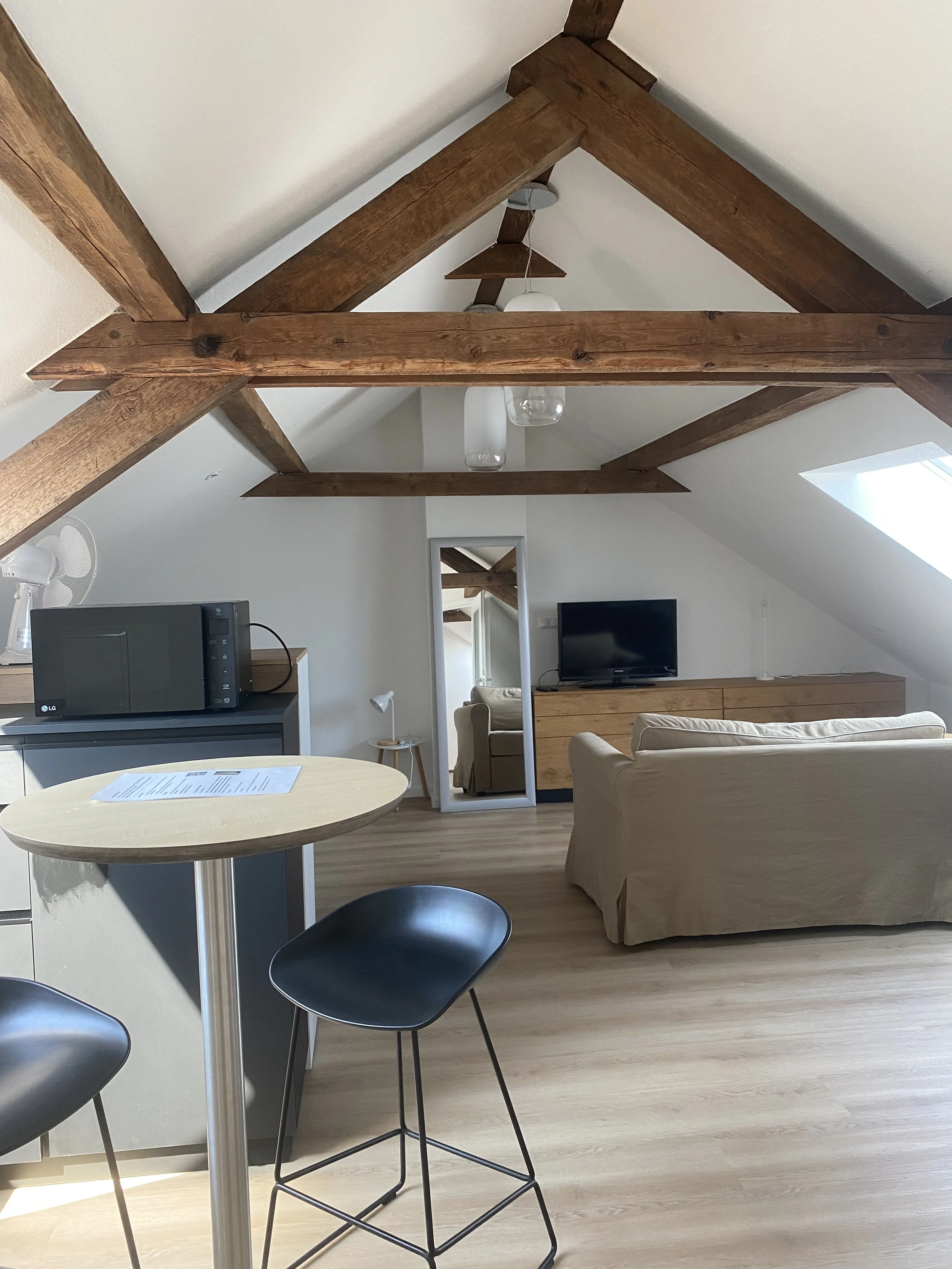 Wohnzimmer mit weißen Wänden, Holzbalkendecke, Sofa, Fernseher, Esstisch, Barhocker, Mikrowelle, und einem Spiegel.