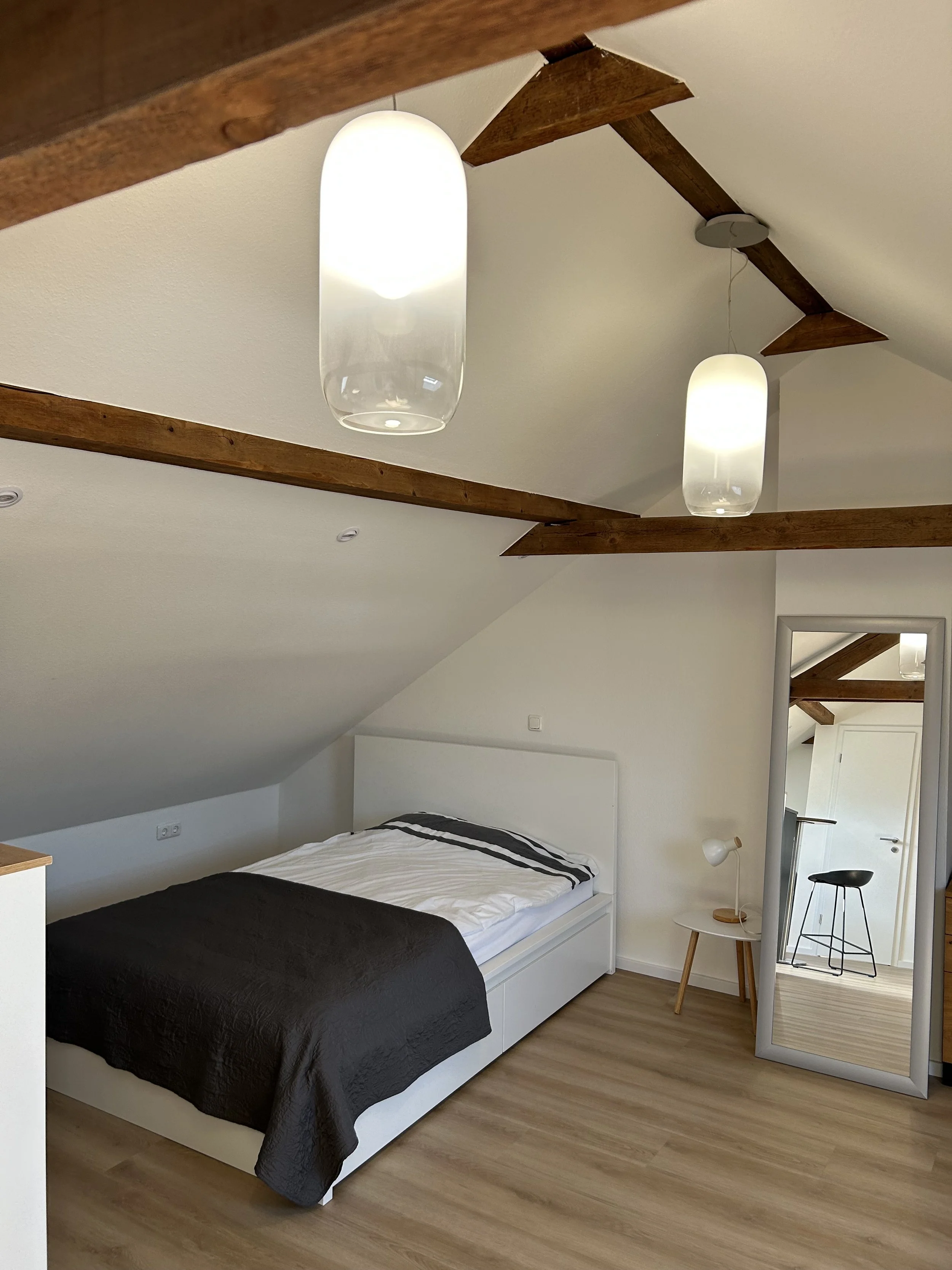 Schlafzimmer mit Dachschräge, Bett mit weißem Rahmen, schwarzen Bettdecke und weißen Kissen, kleiner Nachttisch mit weißer Lampe, großer stehender Spiegel, zwei moderne Deckenleuchten, Holzdecke mit sichtbaren Balken, Holzboden.